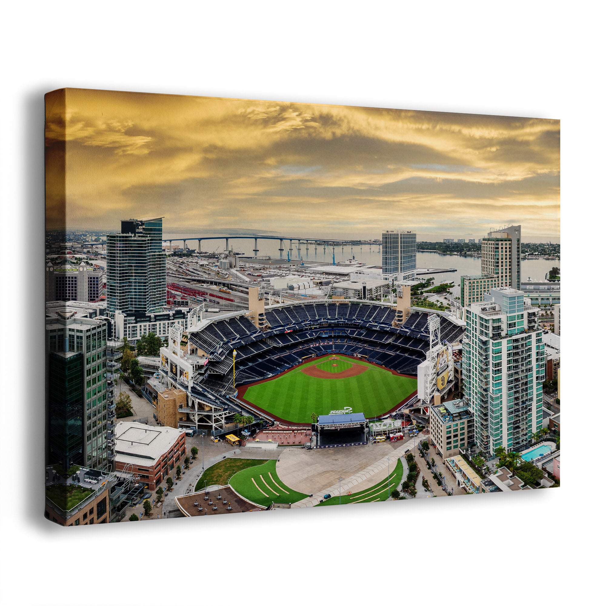 A rectangular canvas wall art print showing an aerial view of the San Diego Padres Petco Park baseball stadium surrounded by city buildings and water under a dramatic golden sky, photographed at an angle against a plain white background.