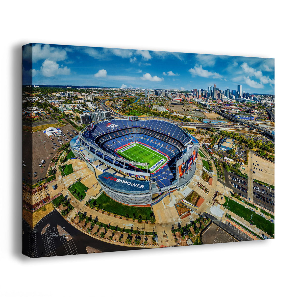 An Empower Field Denver skyline panorama wall art print showing an aerial view of the Broncos' Mile High Stadium with a green field, surrounded by the Denver city skyline under a bright blue sky. The canvas is photographed at an angle against a plain white background.