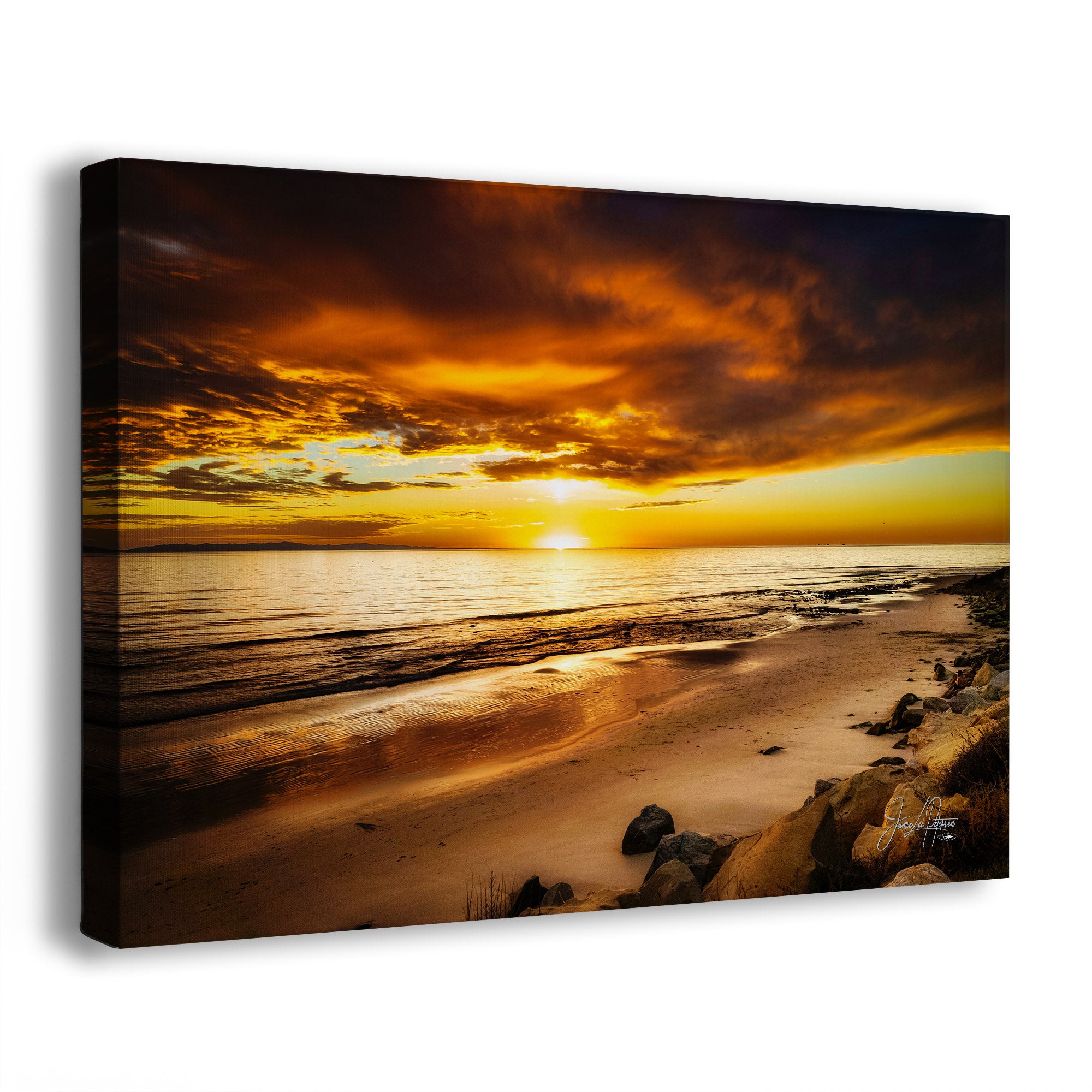 A canvas wall art print showing a dramatic beach sunset with glowing orange and yellow clouds, reflective water, sandy shore, and dark rocks, photographed against a plain white background.