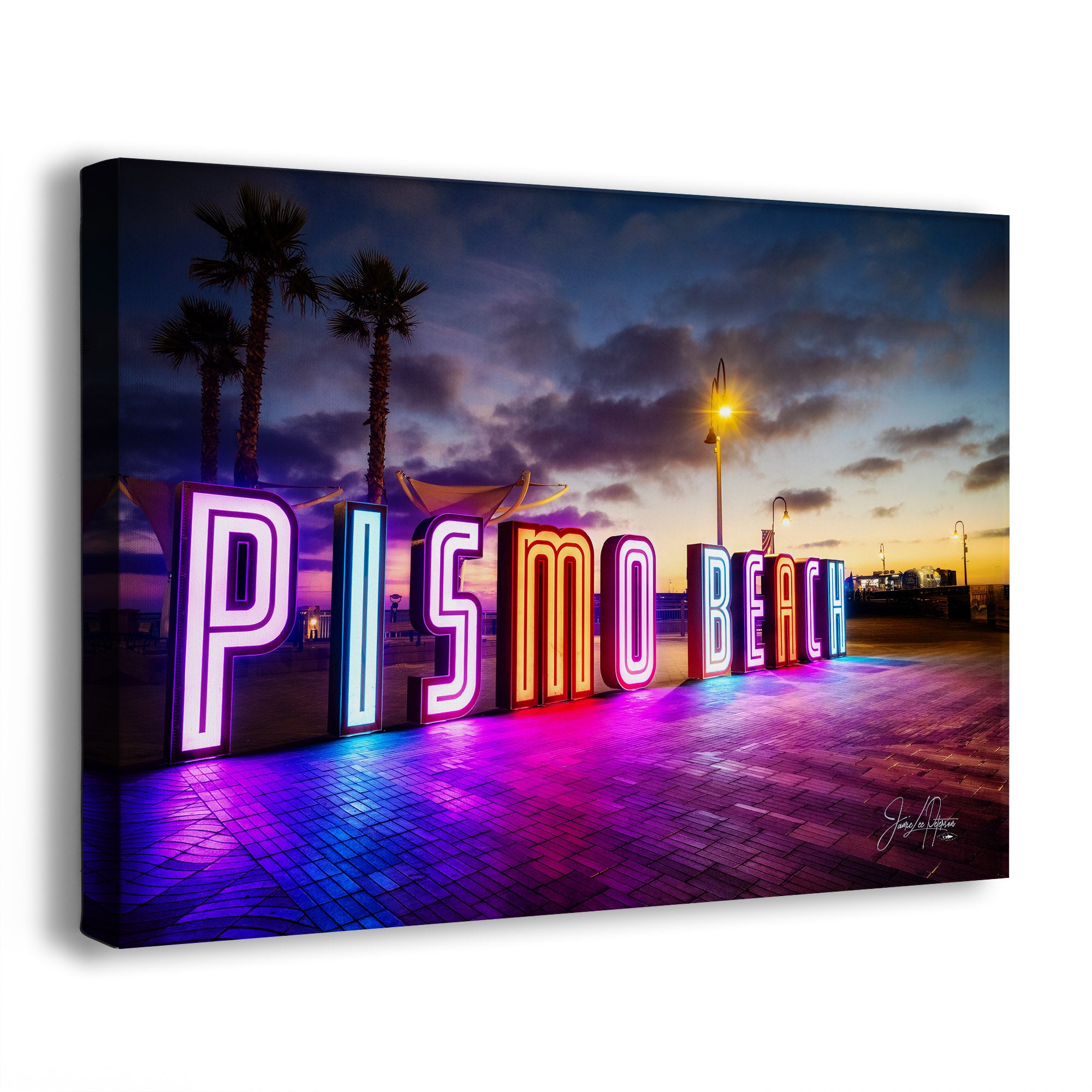 A wall art canvas showing a neon 'Pismo Beach' sign at dusk, with palm trees, a streetlamp, and colorful reflections on the pavement. The canvas is photographed at a slight angle against a plain white background.