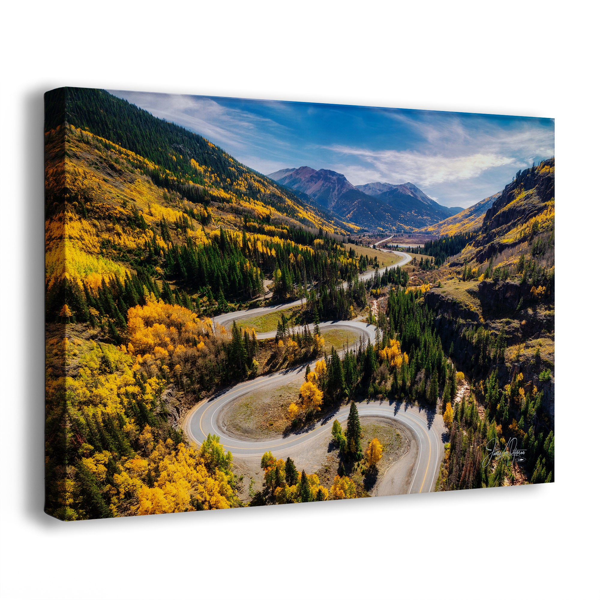 A canvas wall art print showing an aerial Colorado mountain landscape of Million Dollar Highway with a winding road, yellow autumn trees, evergreen forest, and blue sky, photographed against a light gray background.