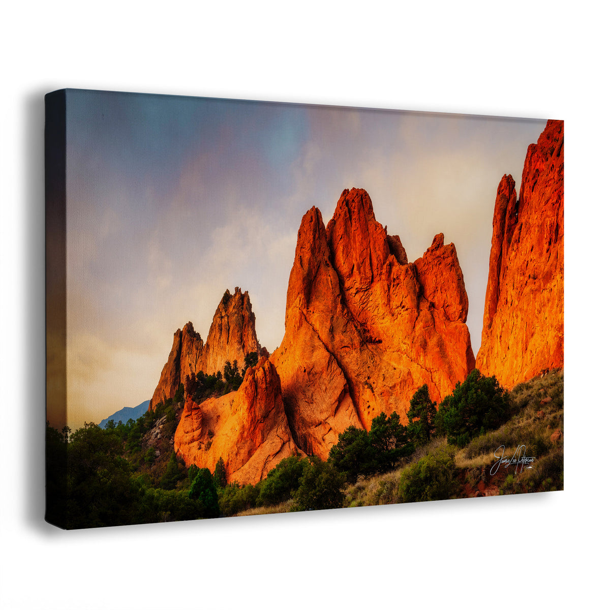 A canvas wall art print showing a dramatic desert rock formation scene at sunset, photographed at a slight angle against a plain white background. The artwork depicts glowing orange-red rock spires with green trees and a cloudy sky.