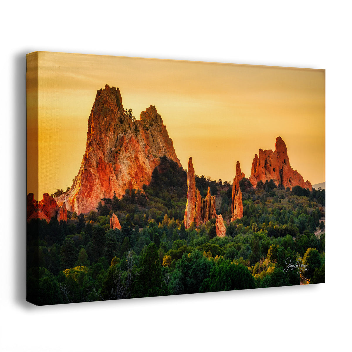 A rectangular canvas wall art print showing a dramatic desert rock formation at Garden of the Gods in Colorado at sunset, with a glowing orange and yellow sky, dark red rock spires, and green trees in the foreground. The canvas is photographed at a slight angle on a clean white background.