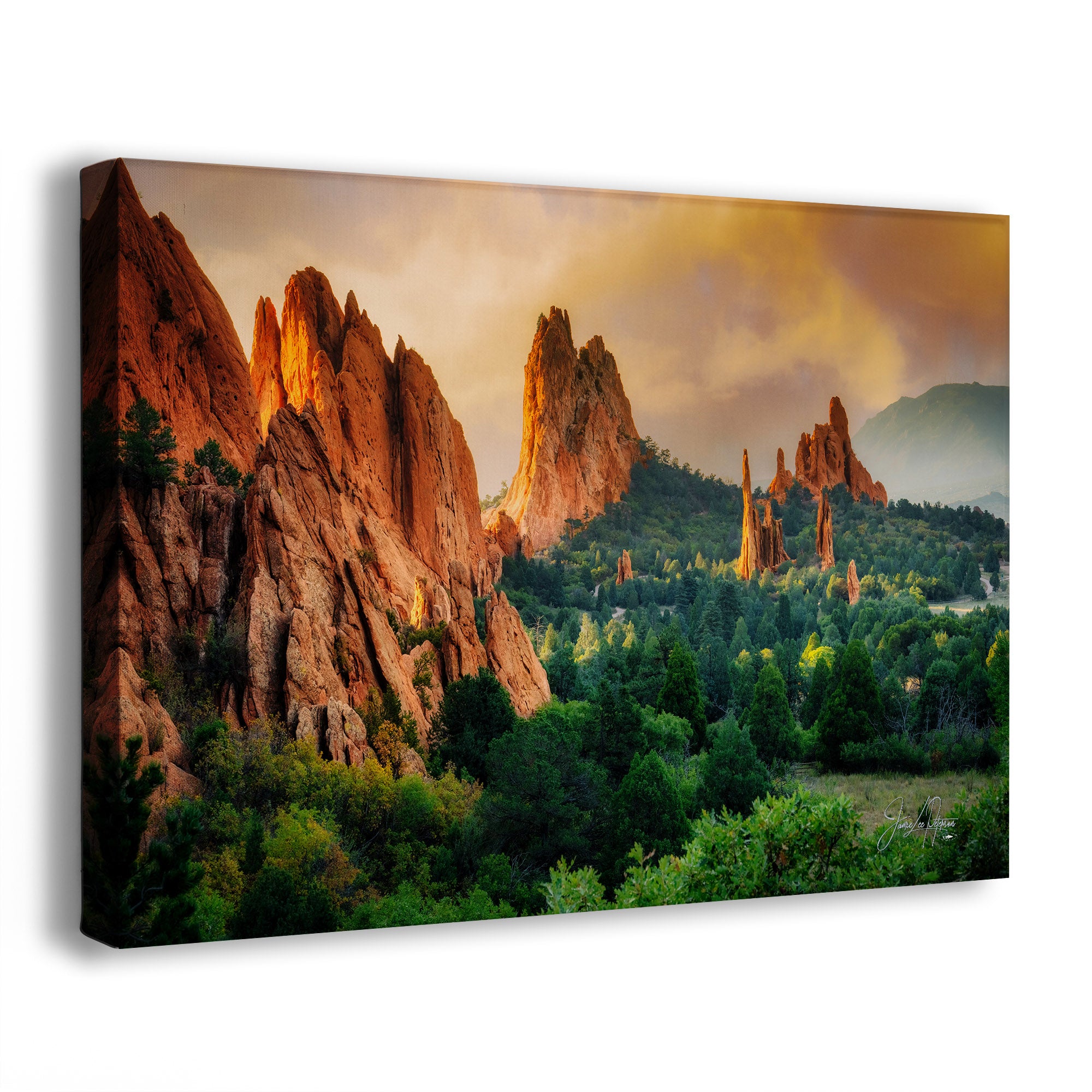 A canvas wall art print showing a dramatic natural landscape from Garden of the Gods, Colorado, with tall orange-red rock spires and green trees under a warm golden sky, photographed at an angle against a light gray background.