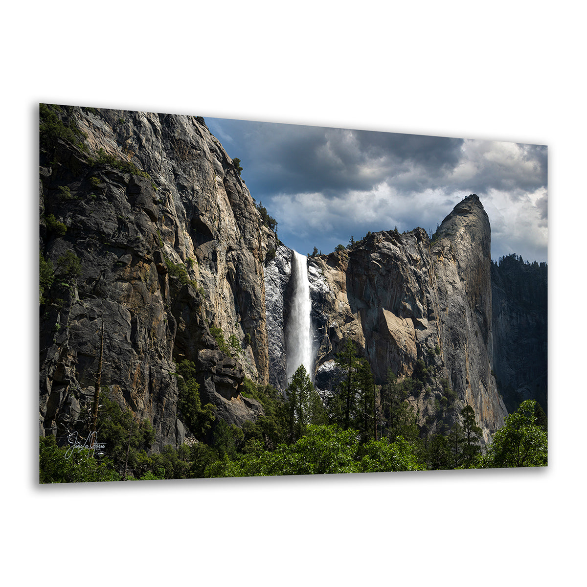 A rectangular Yosemite wall art decor print showing a dramatic natural landscape with the tall Bridalveil waterfall between tall rocky cliffs, green trees in the foreground, and a cloudy blue sky. The print is photographed against a plain white background with a slight perspective angle.