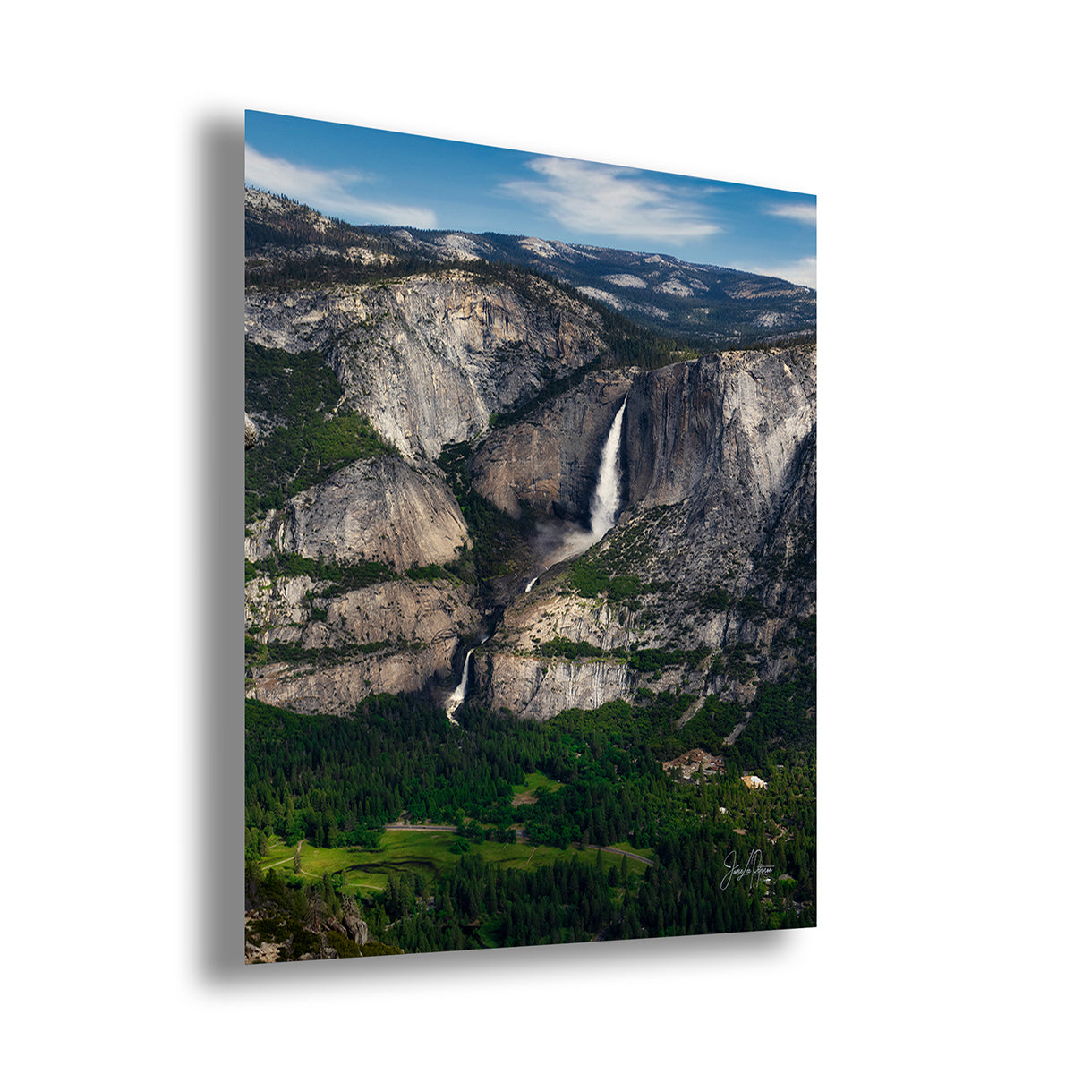A vertical wall art decor print of Yosemite Falls depicting a dramatic mountain landscape with a tall waterfall, rocky cliffs, green forest, and blue sky, shown at an angle against a white background.