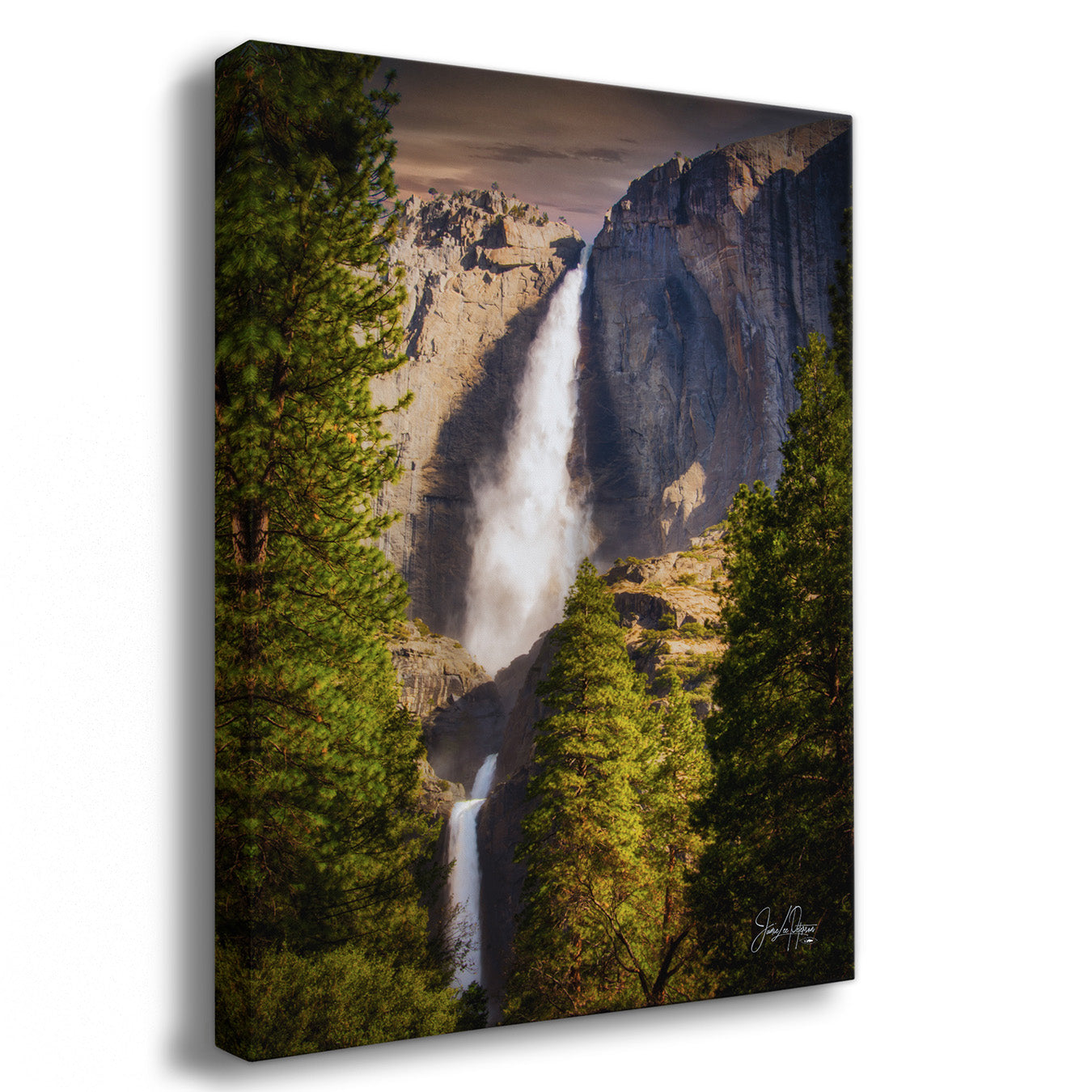 A vertical canvas wall art print depicting a dramatic waterfall cascading down a rocky cliff, framed by tall green pine trees in the foreground. The canvas is shown at a slight angle against a plain white background.