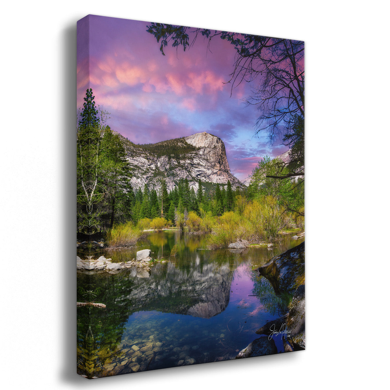 A vertical canvas wall art print showing a mountain reflected in a calm lake, with pine trees, yellow-green shrubs, and a dramatic pink and purple sky. The canvas is photographed at a slight angle against a clean white background.
