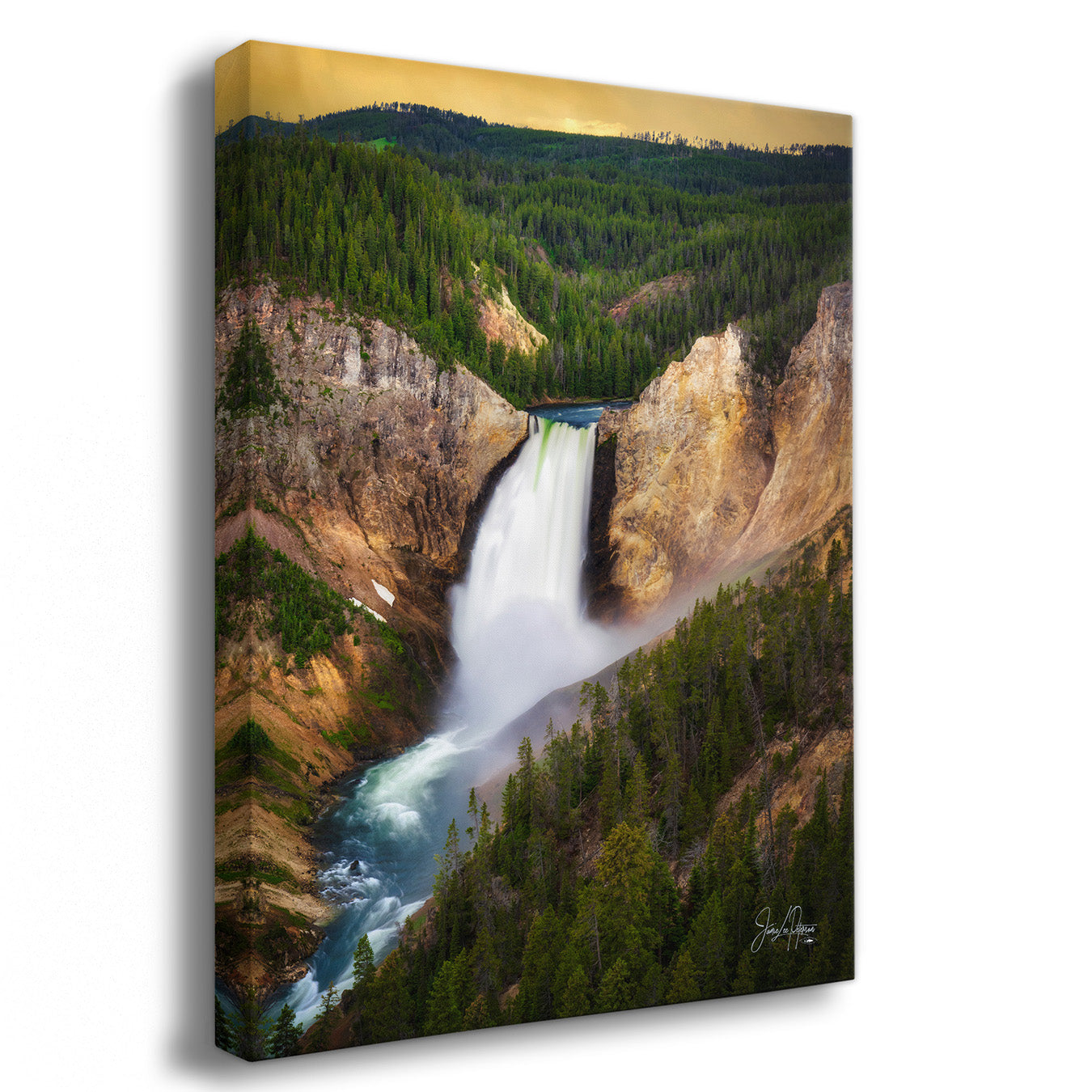 A vertical canvas wall art print depicting a dramatic waterfall cascading down steep rocky cliffs into a river, surrounded by dense evergreen forest under a warm golden sky. The canvas is shown at a slight angle with a drop shadow on a white background.