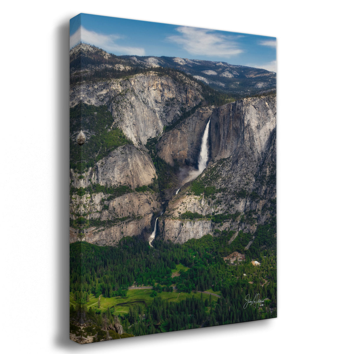 A Yosemite Falls mountain art print depicting a dramatic mountain landscape from Yosemite National Park with the tall Yosemite Falls, rocky cliffs, and dense green forest under a blue sky, shown at a slight angle against a white background.