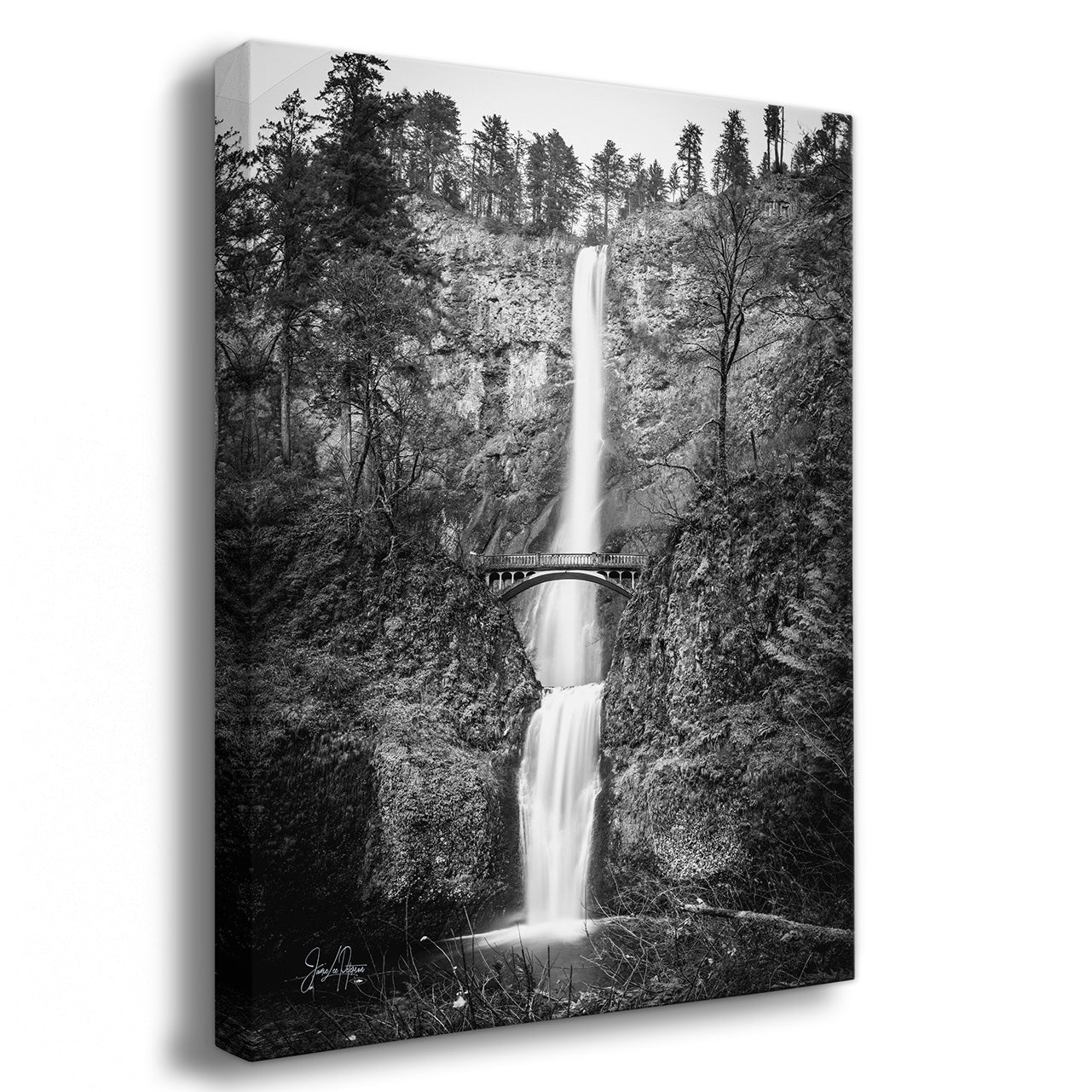 A vertical black and white canvas wall art print depicting a tall waterfall cascading down a rocky cliff with trees above and a small bridge across the middle, shown at a slight angle with a drop shadow on a white background.