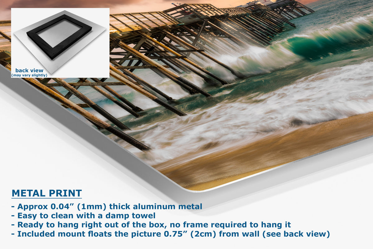 A corner view of a metal wall art print showing a beach scene with ocean waves, wooden pier structures, and sandy shore, presented at an angle with product text and a small back-view inset on a white background.