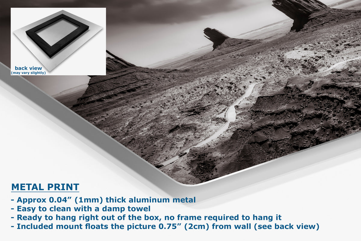 A corner view of an aluminum metal wall art print showing a black-and-white desert landscape with a winding path, presented at an angle with product text and a small back-view inset. The text says 'METAL PRINT' and describes the print.