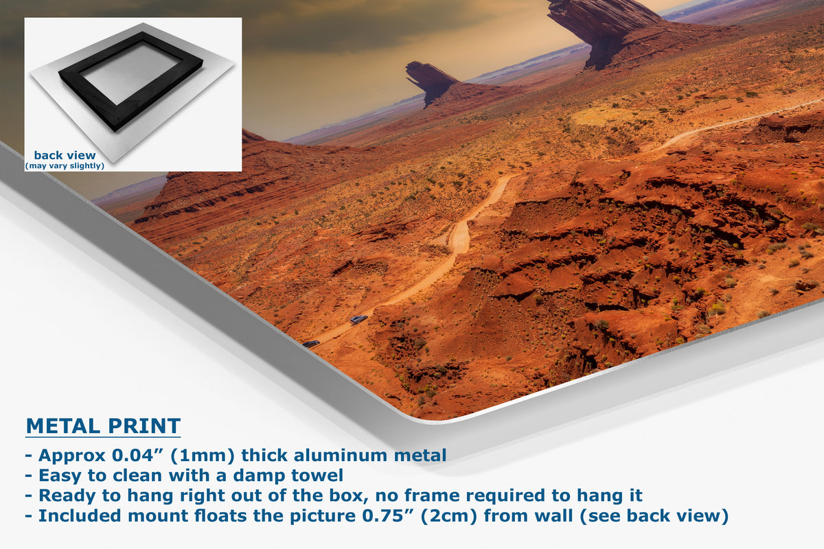 A corner view of a metal wall art print showing an aerial desert landscape with reddish-brown rock formations and a winding road, presented at an angle with a small inset showing the back view. Text on the image describes the product.