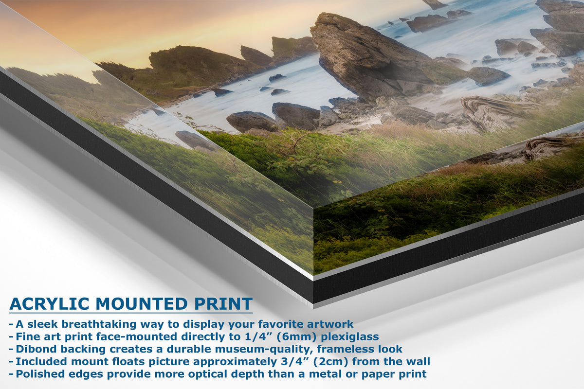 Acrylic mounted art print of a coastal landscape with rocky cliffs, green foreground, and ocean waves under a warm sunset sky. The image includes product feature text at the bottom describing the acrylic frame, plexiglass, mounting, and frameless look.