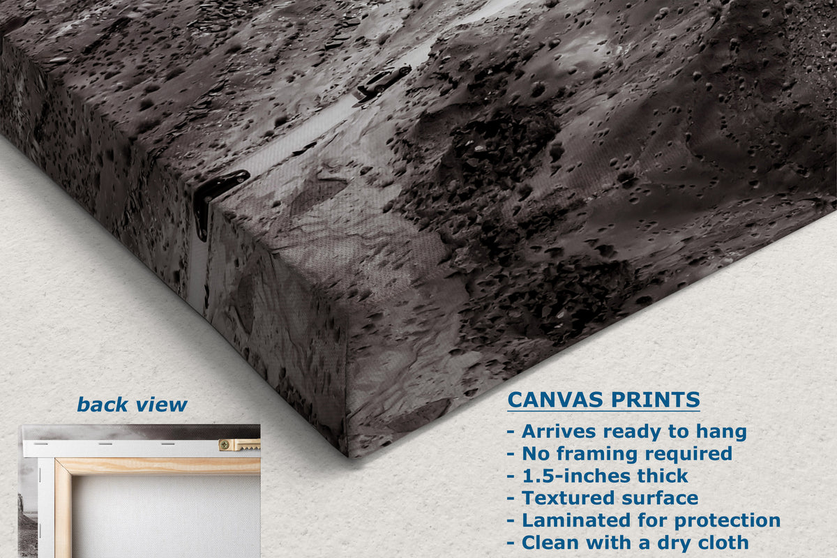 A corner view of a canvas wall art print shown at an angle, displaying a textured black and gray abstract landscape design. The image includes a text overlay detailing product features ('CANVAS PRINTS') and a small inset showing the back view with a wooden frame.