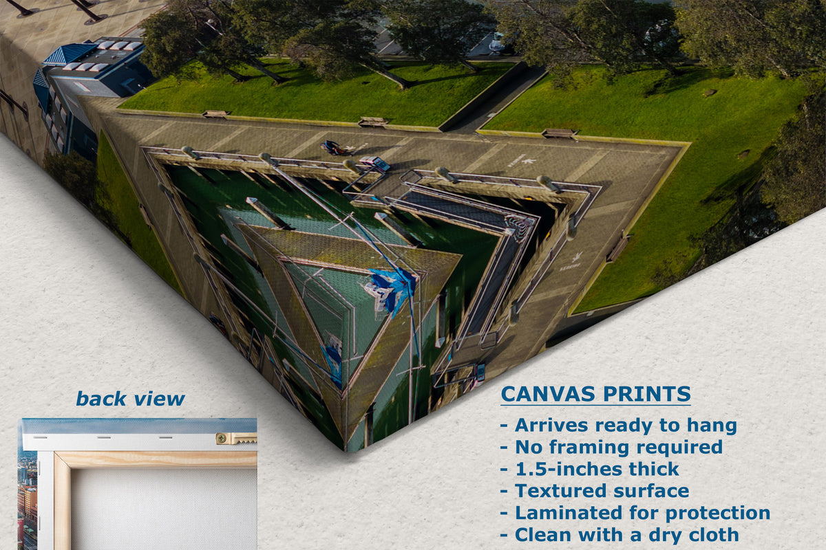 A corner view of a canvas wall art print shown at an angle with a text panel describing features and a small inset labeled back view. The artwork depicts an aerial architectural scene with green grass, gray pathways, and dark blue geometric structures.