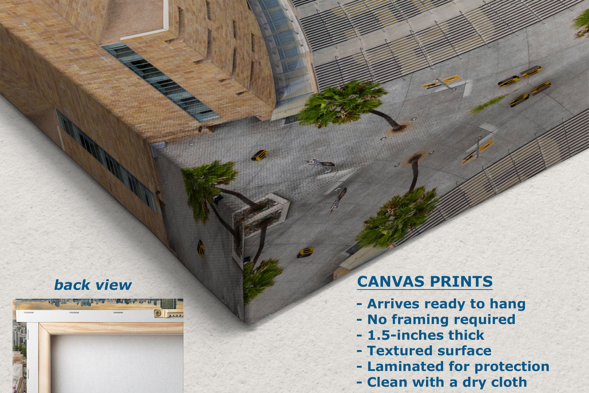 A printed canvas wall art print shown at an angle, displaying a detailed architectural scene of a modern building with a plaza and palm trees. The image includes text overlays detailing product features for canvas prints and a small inset showing the back view of the print.