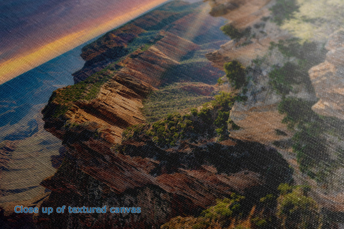 Close-up of a textured canvas print depicting a canyon landscape with layered reddish-brown rock formations and green vegetation under a warm sunset sky. Blue text in the lower-left corner reads 'Close up of textured canvas'.