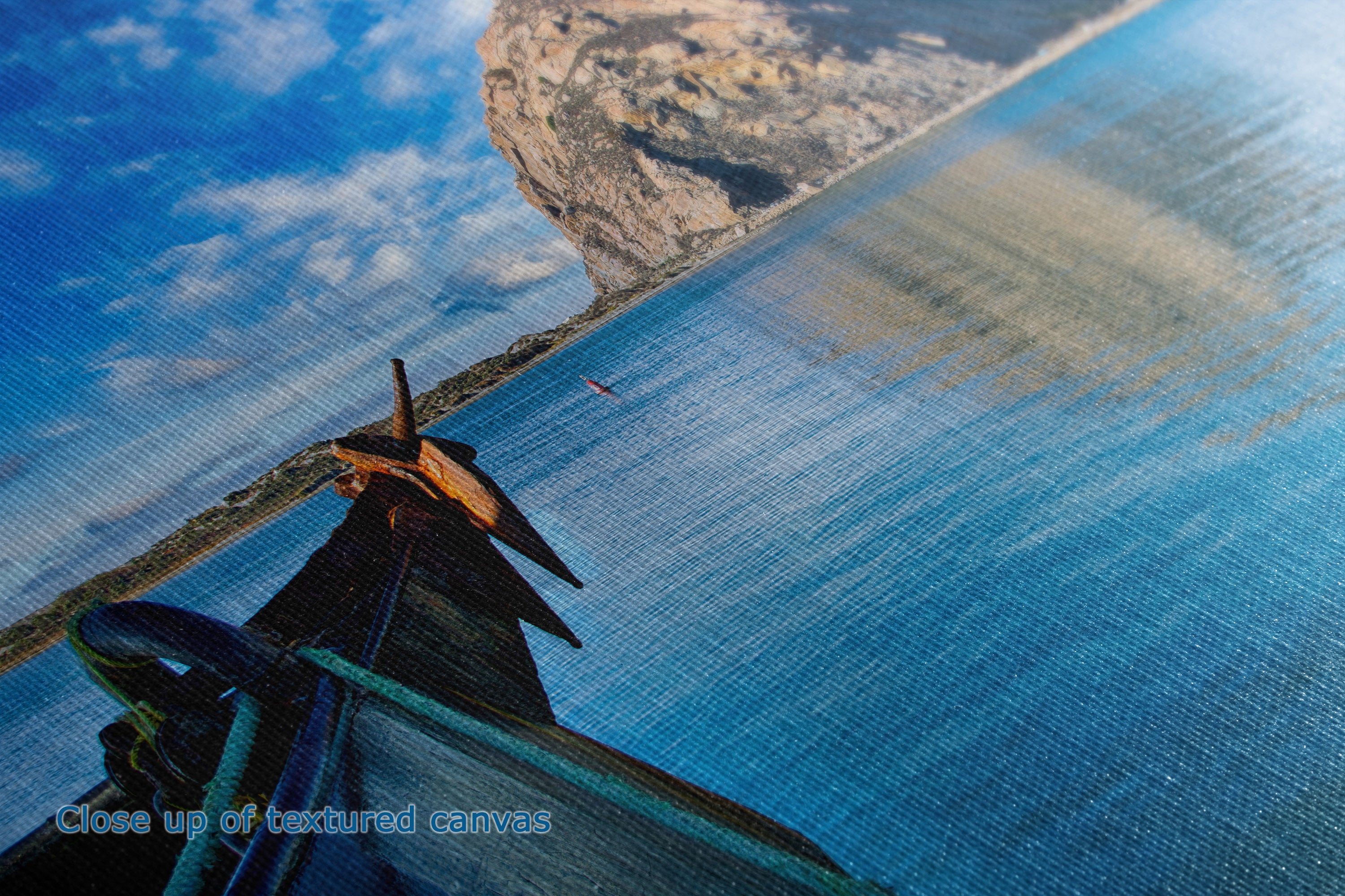 Close-up of a large textured canvas print depicting a coastal landscape with blue water, rocky cliffs, and sky, with a dark sailboat in the lower left corner.