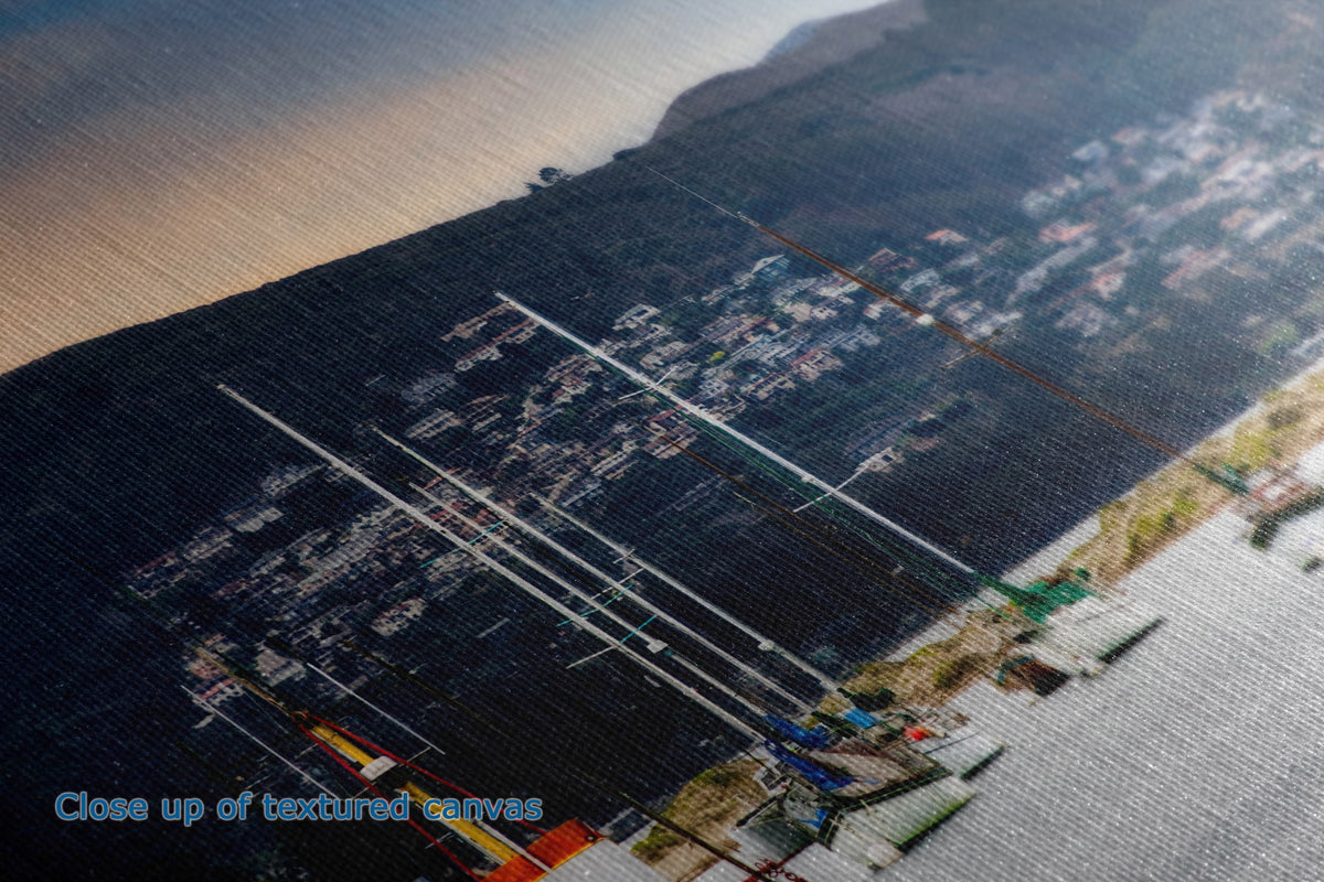 A close-up textured shot of a canvas print depicting an aerial view of a coastal harbor with sailboats, a dark hillside town, and water, with a blue text overlay reading 'Close up of textured canvases'.
