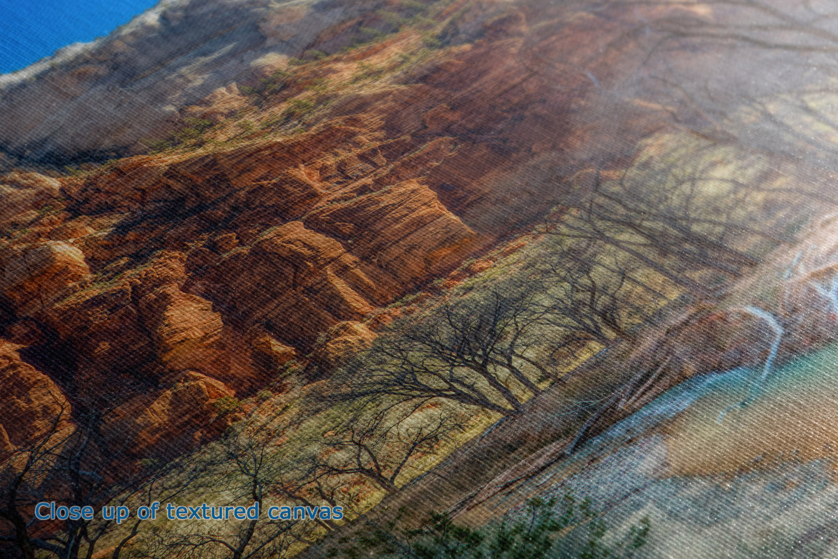 Close-up of the detail and texture of a canvas print depicting a desert canyon landscape with reddish-brown rock formations, sparse leafless trees, and a winding river under a blue sky.
