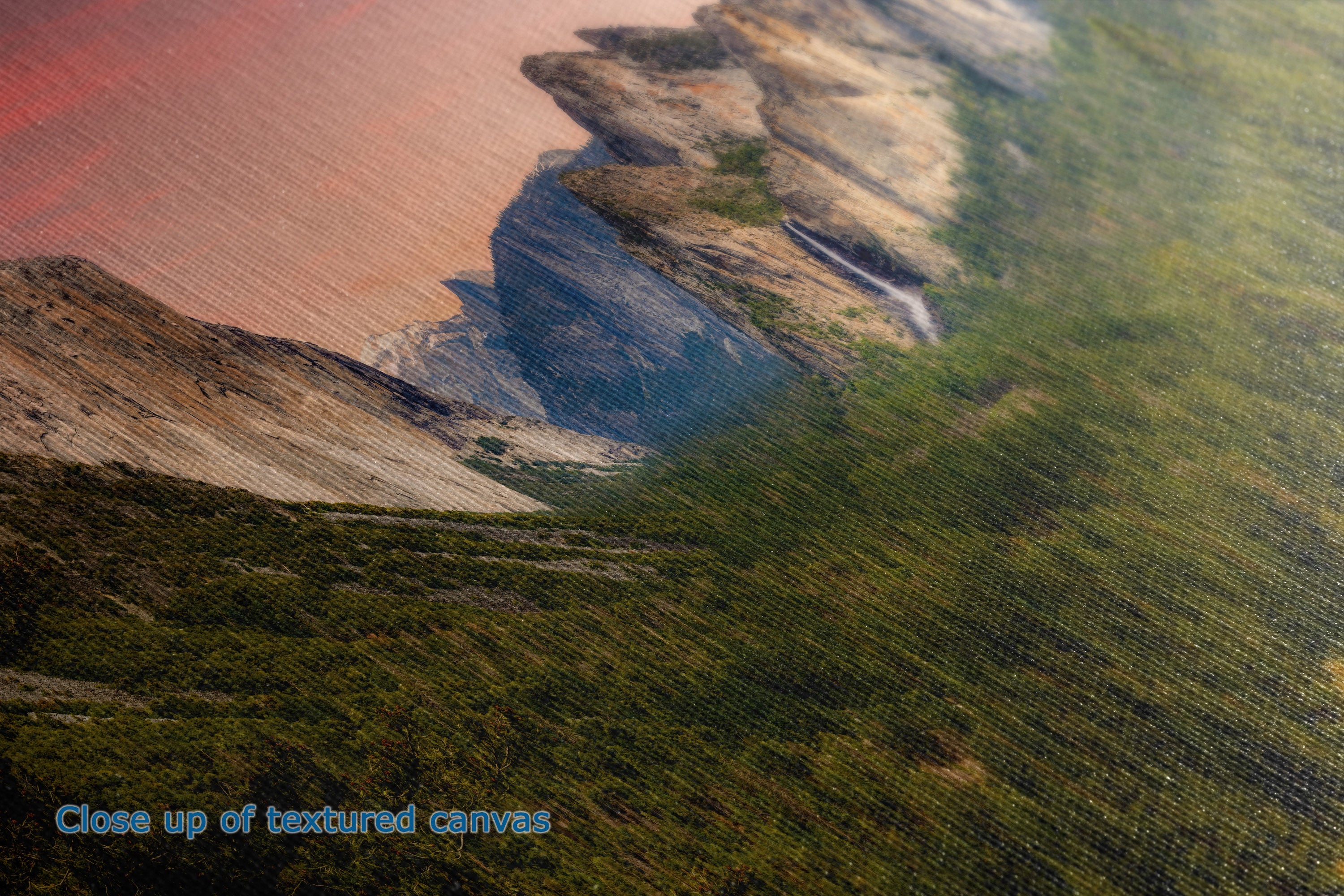 Close-up of a textured canvas print depicting a coastal landscape with reddish-brown cliffs, blue water, and green vegetation, with the text 'Close up of textured canvases' in the bottom-left corner.