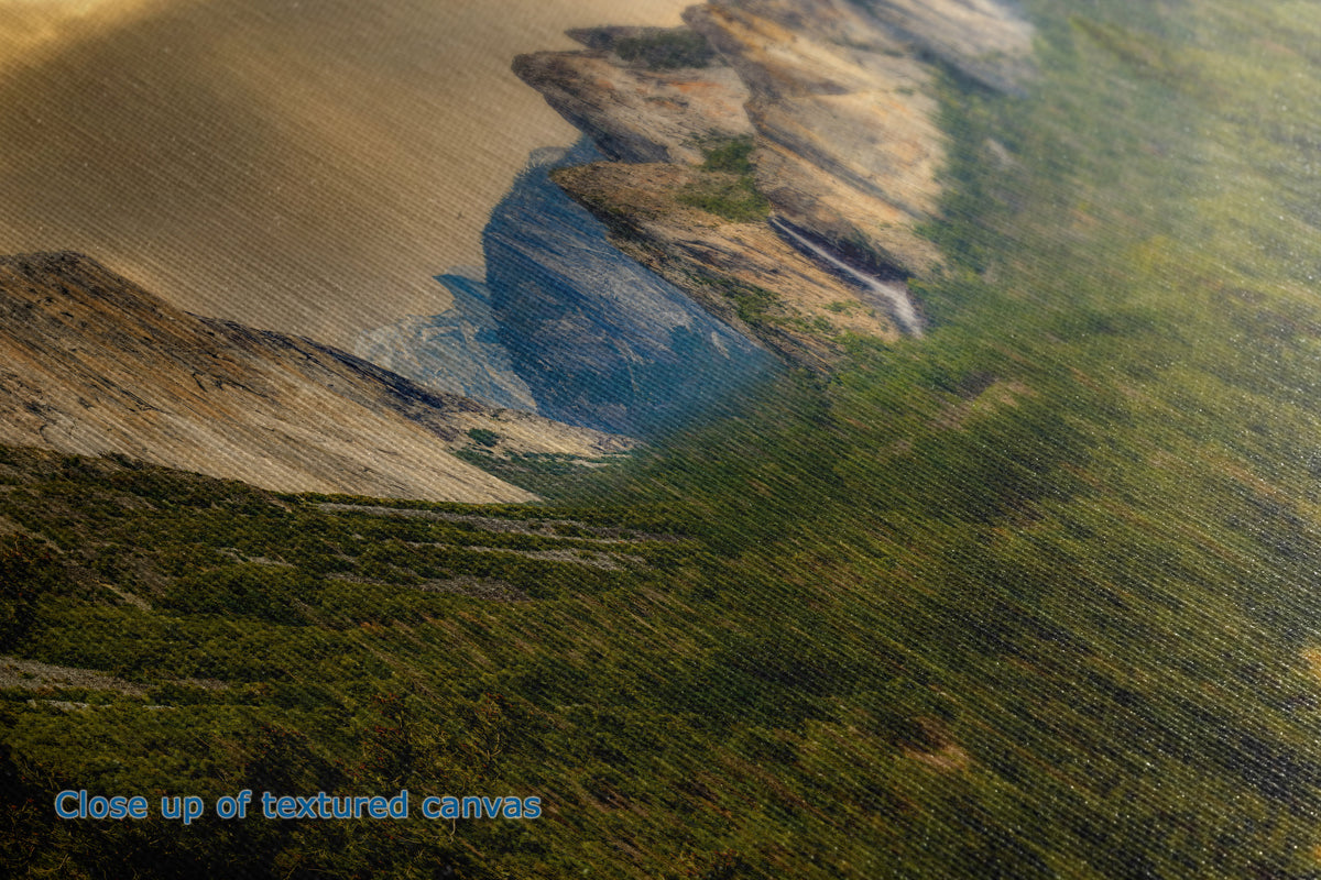Close-up of a textured canvas print depicting a dramatic landscape with steep cliffs, a blue river, and green vegetation. Blue text in the lower-left corner reads 'Close up of textured canvases'.