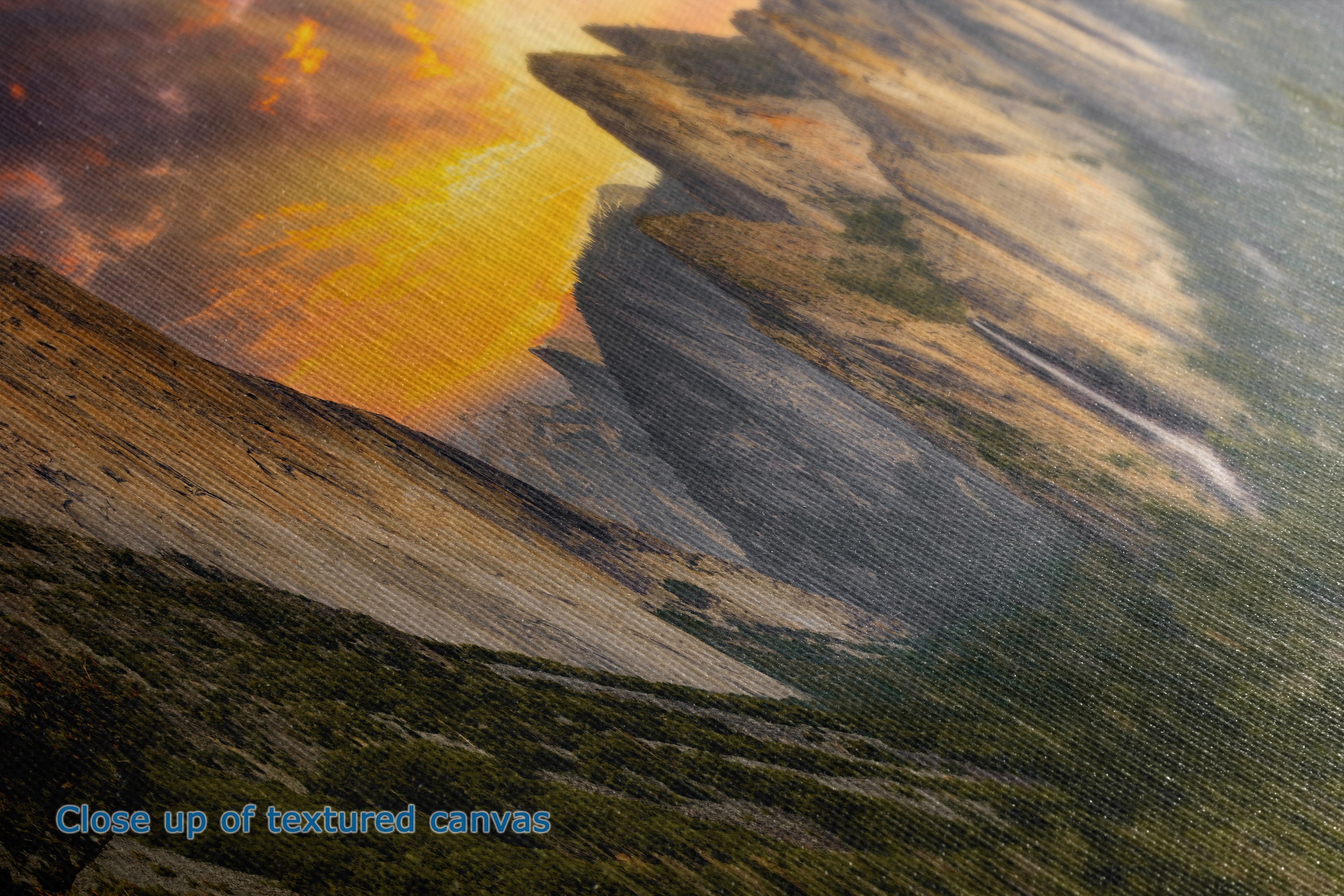 Close-up of the detail and texture of a  canvas print depicting a dramatic mountain landscape at sunset, with glowing orange and yellow clouds, dark rocky cliffs, and green-brown terrain. Blue text in the lower-left corner reads 'Close up of textured canvas'.