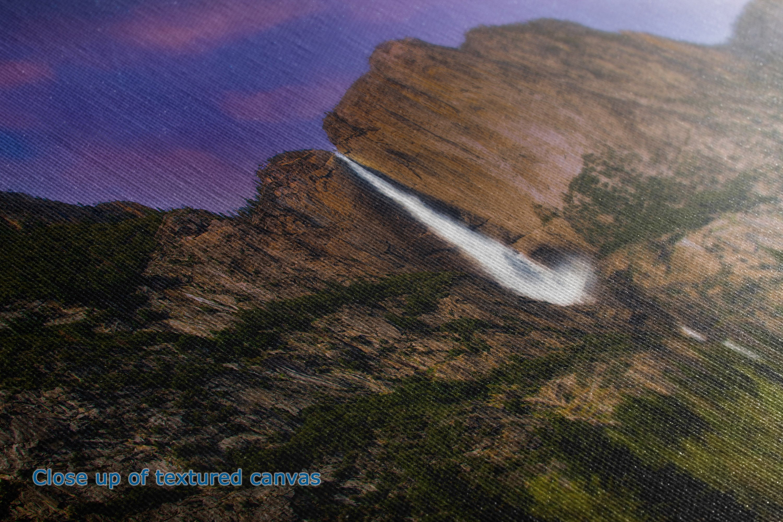 Close-up of a textured canvas print depicting a dramatic mountain landscape with a waterfall under a purple and pink sky, with blue text in the lower-left corner reading 'Close up of textured canvas'.