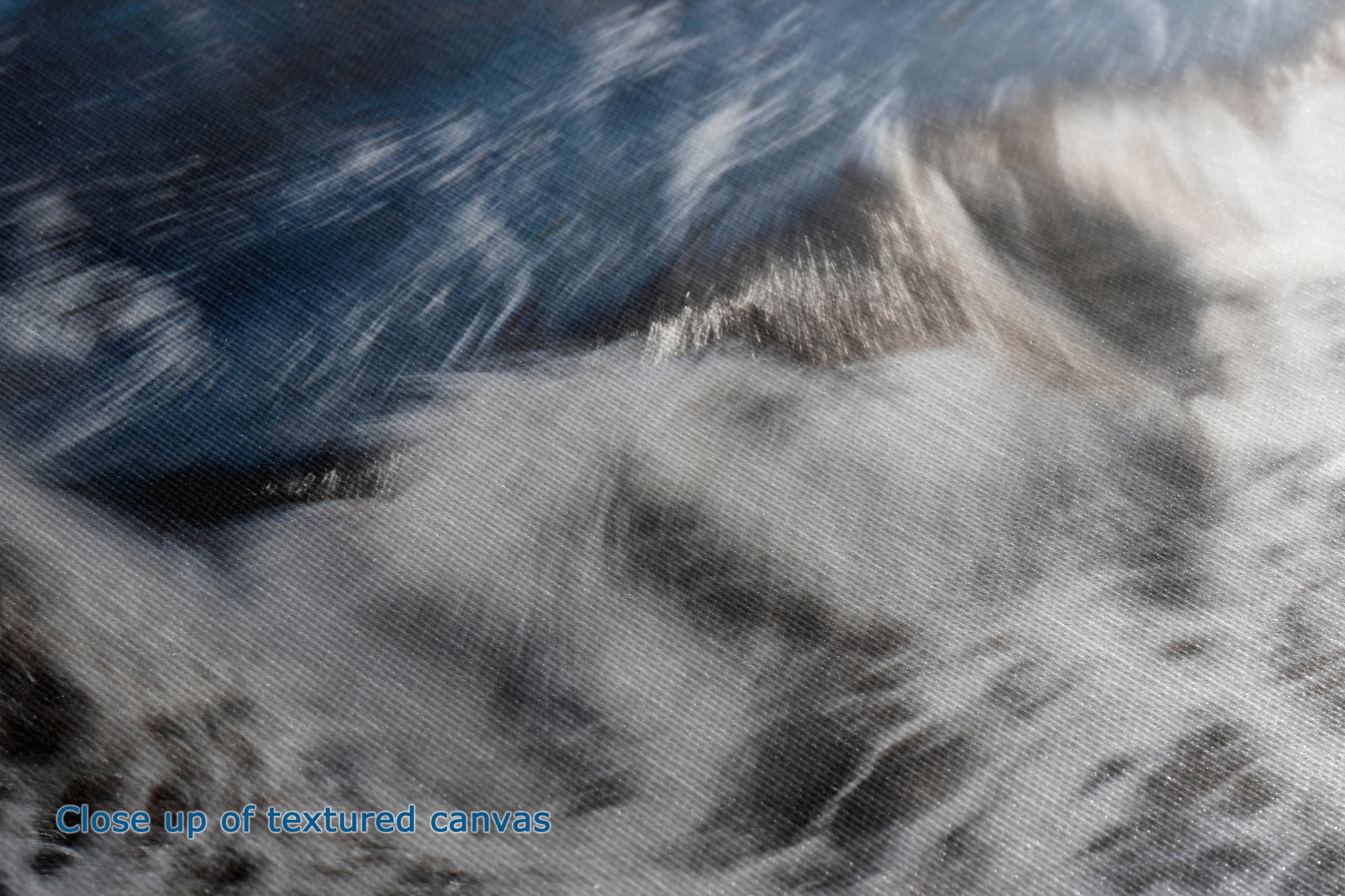 Close-up of a textured canvas print depicting a dramatic scene of white waves crashing against a dark blue shore, with light blue text in the bottom-left corner reading 'Close up of textured canvas'.