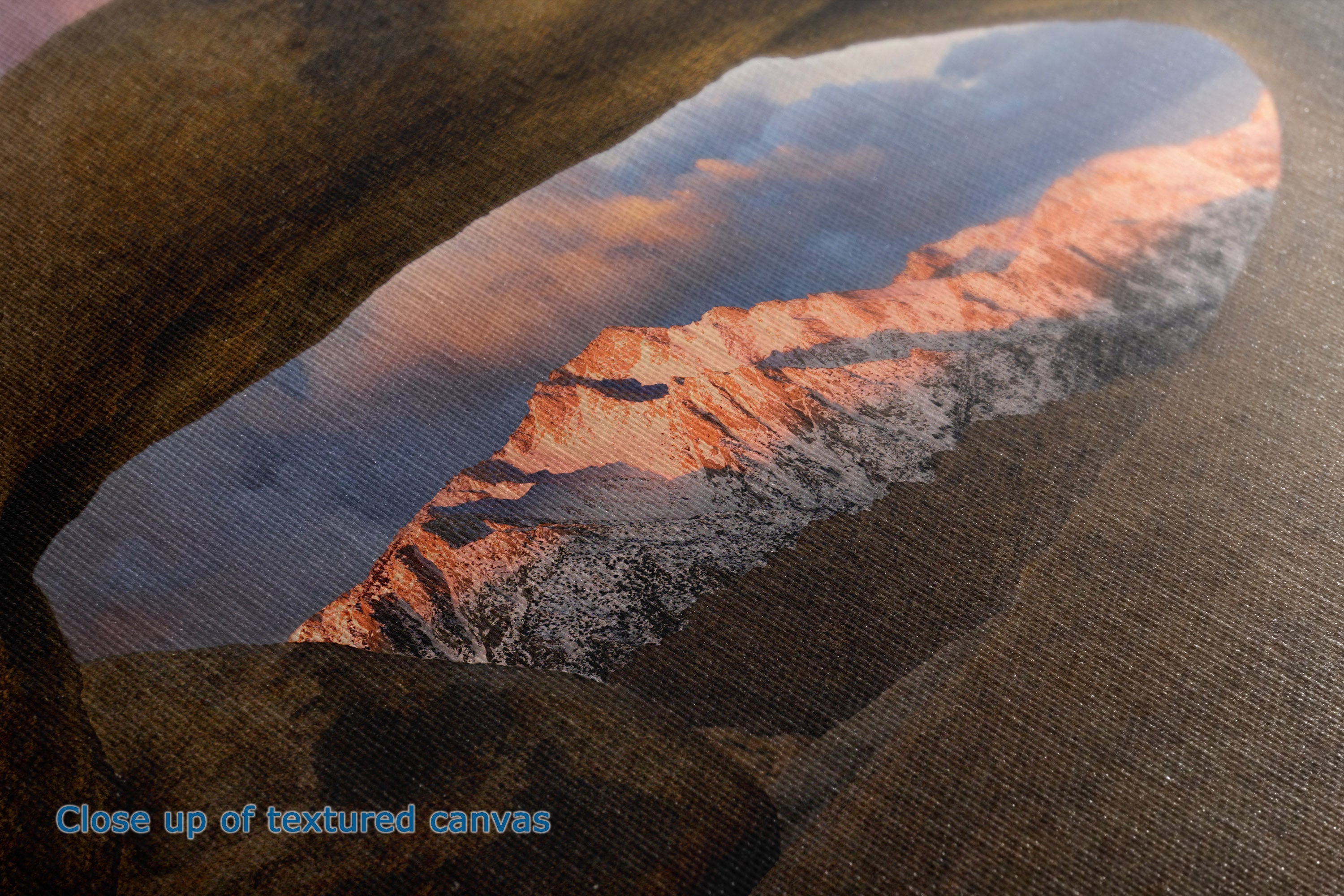 Close-up of a textured canvas print featuring a mountain landscape with snow-capped peaks under a cloudy sky, with blue text in the lower-left corner reading 'Close up of textured canvas'.