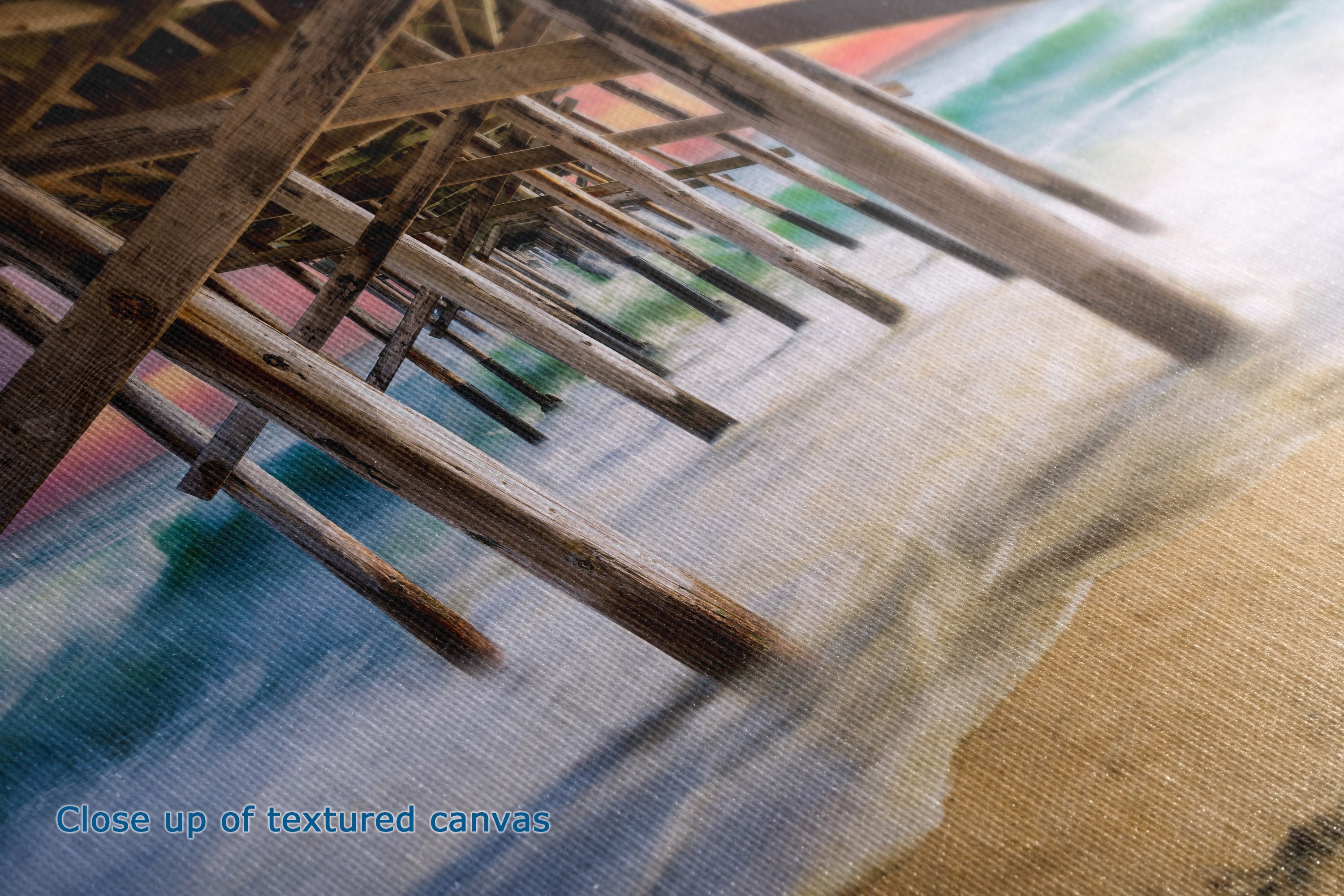 Close-up of a textured canvas print showing a beach scene with wooden pier supports in the foreground and blue, green, beige, and brown tones. Text in the lower-left corner reads 'Close up of textured canvas'.