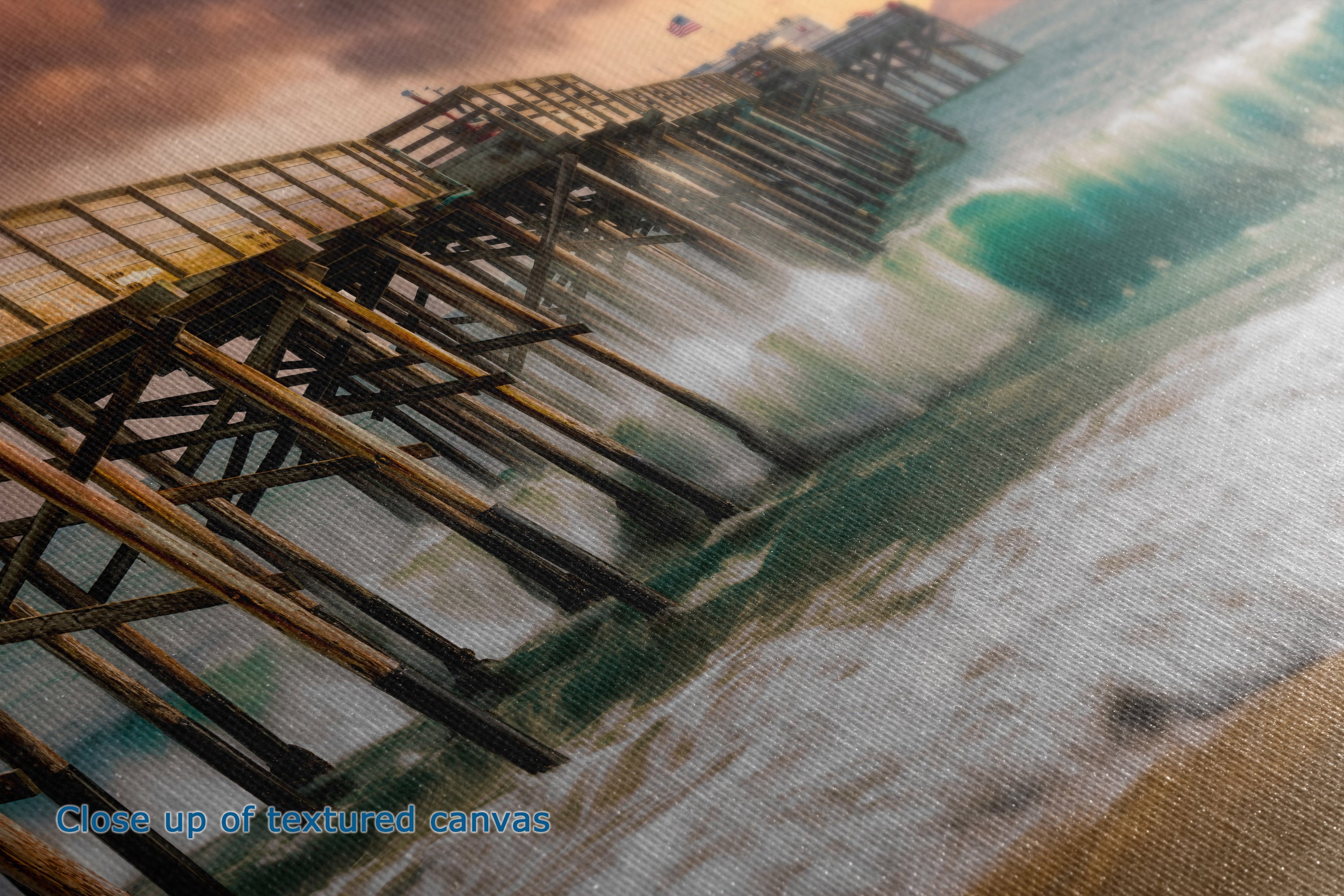 Close-up of a textured canvas print depicting a wooden pier extending into ocean waves at sunset, with a small blue text overlay reading 'Close up of textured canvas.'.