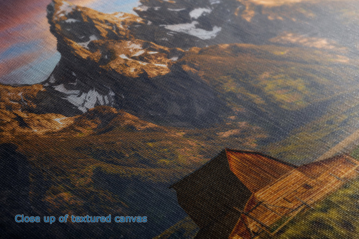 Close-up of a textured canvas print depicting a mountain landscape with snow patches, warm golden light, and a cabin roof in the lower right corner.