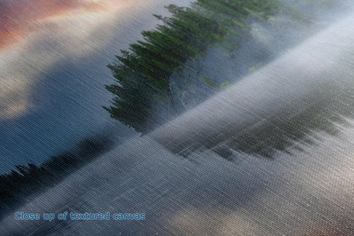 A close-up view of the texture and detail of a canvas print that depicts a misty forest landscape with dark green pine trees, gray-blue fog, and warm orange sky tones. 
