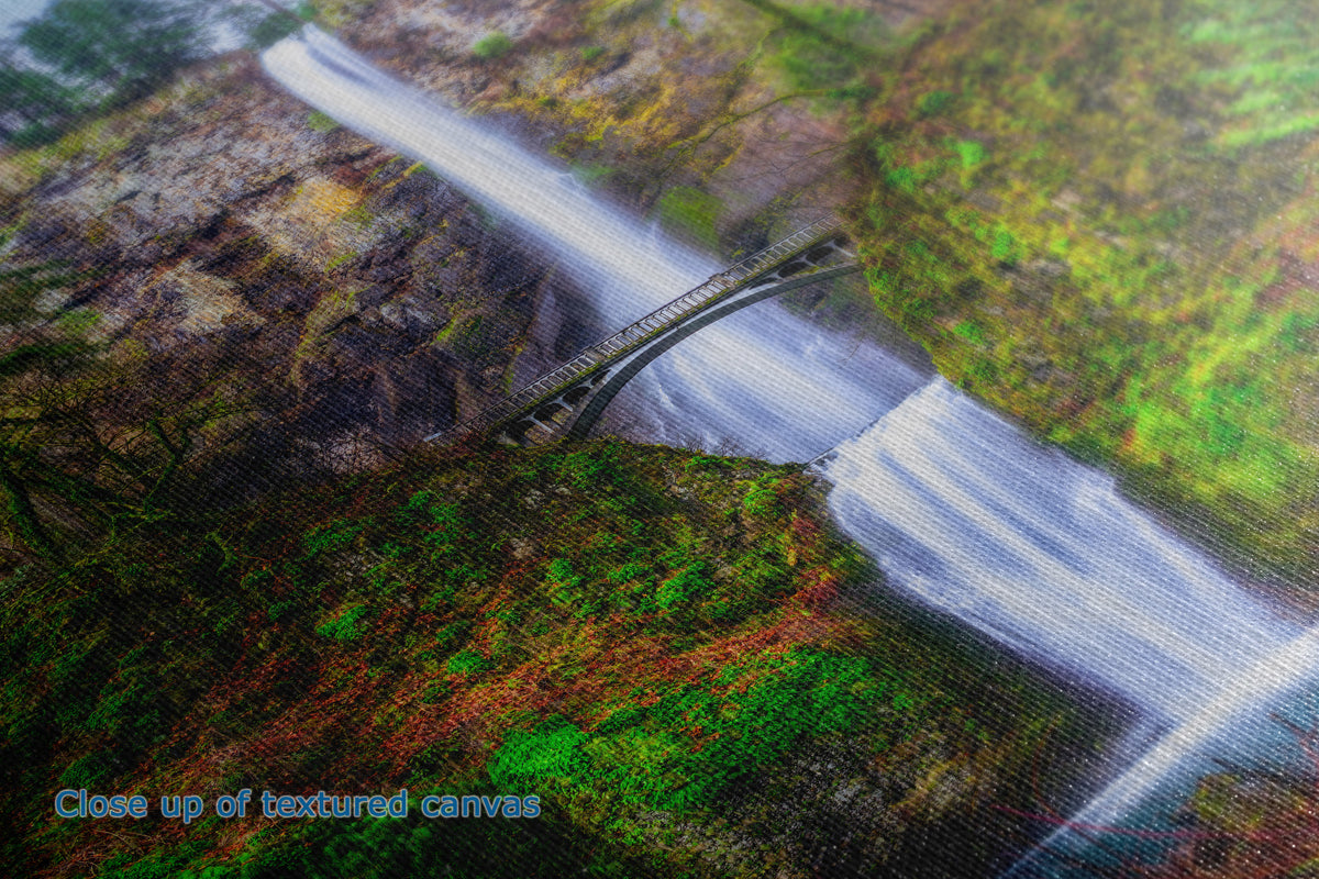 A close-up of a textured canvas print depicting a scenic landscape with a waterfall, a small bridge, and lush green and brown foliage.