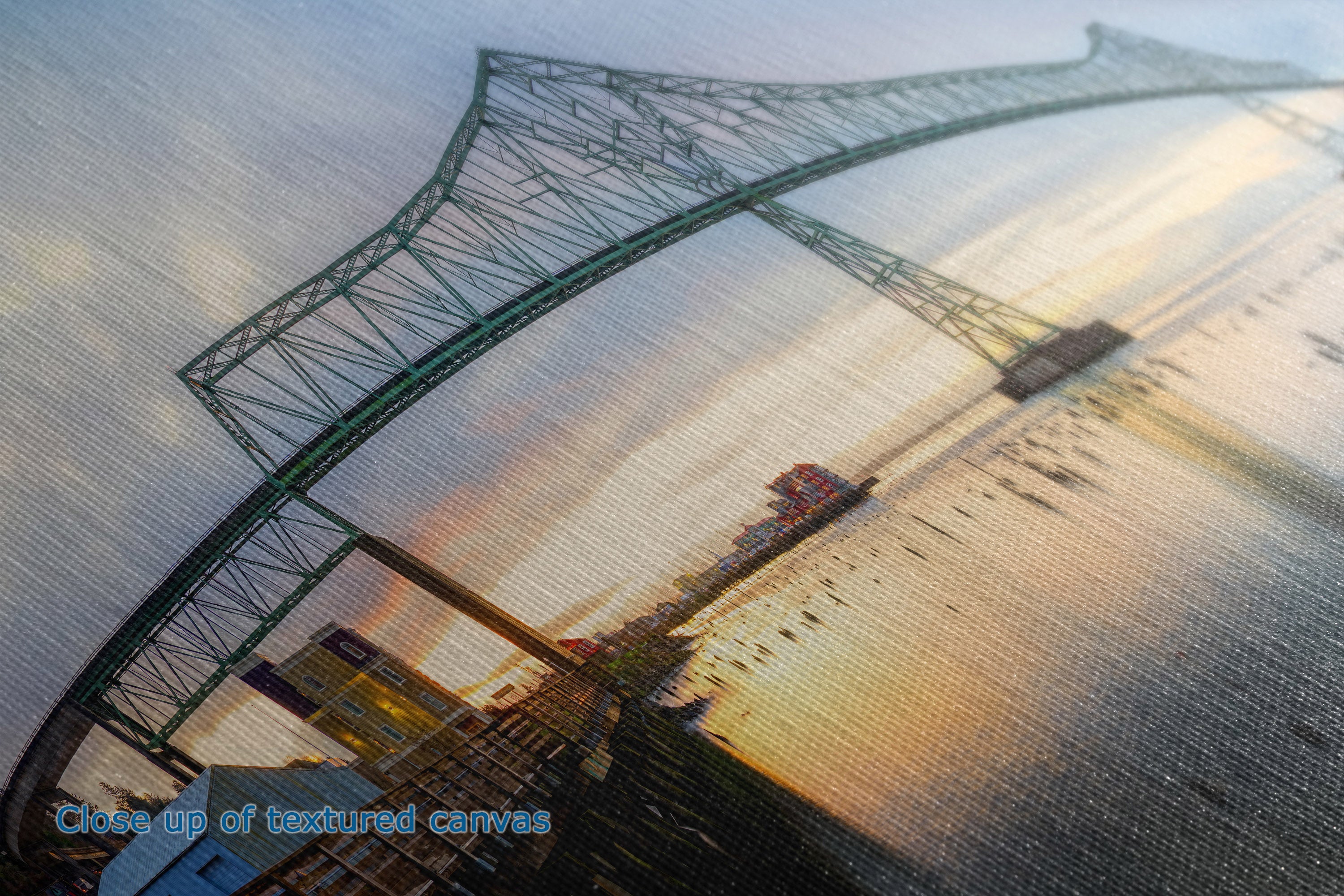 Close-up of a textured canvas print depicting a large suspension bridge over water at sunset, with warm golden light and cool blue-gray tones.