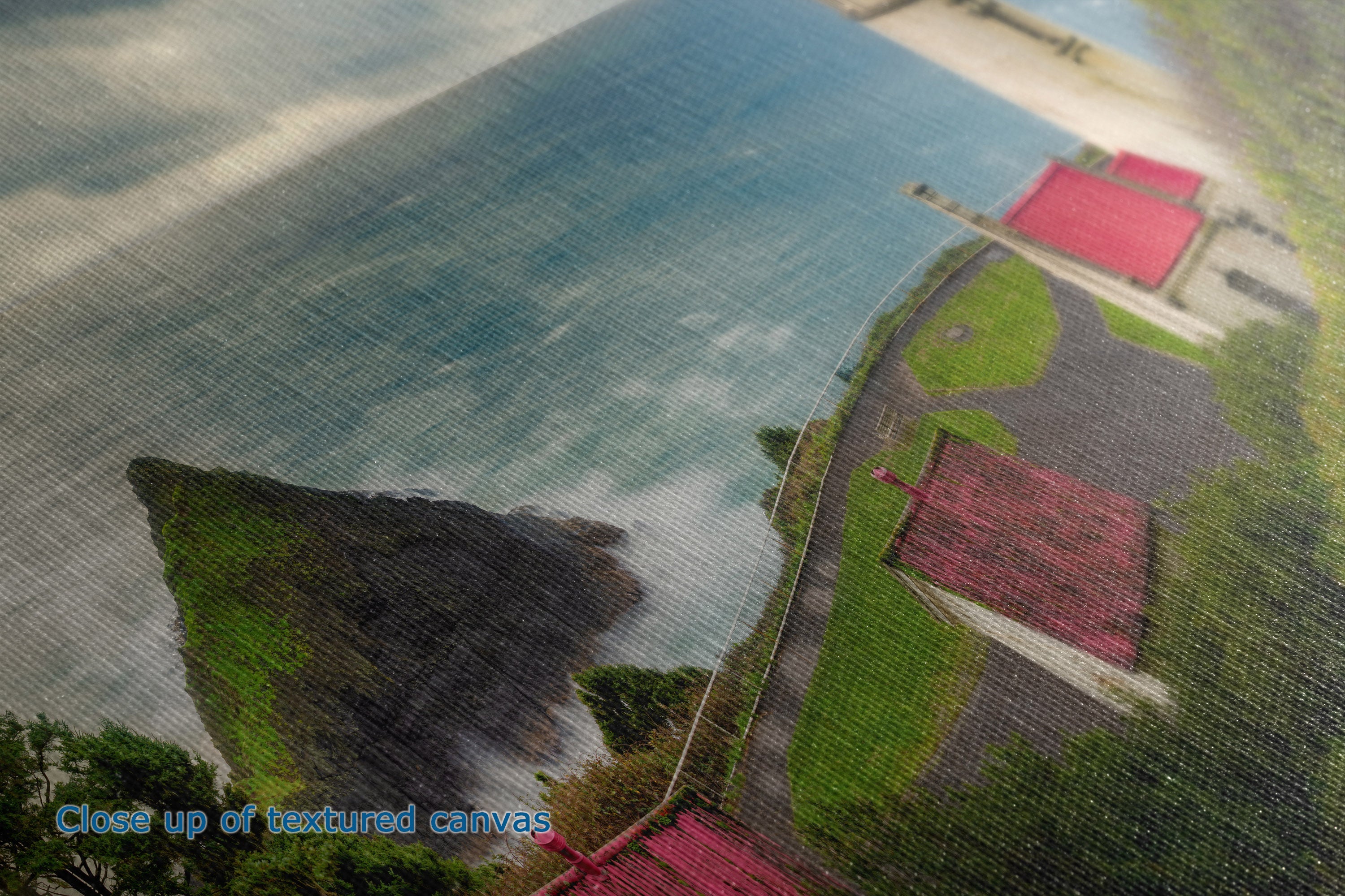 Close-up of a textured canvas print depicting a coastal landscape with blue water, green cliffs, and red-roofed buildings, with the text 'Close up of textured canvas' in the lower-left corner.