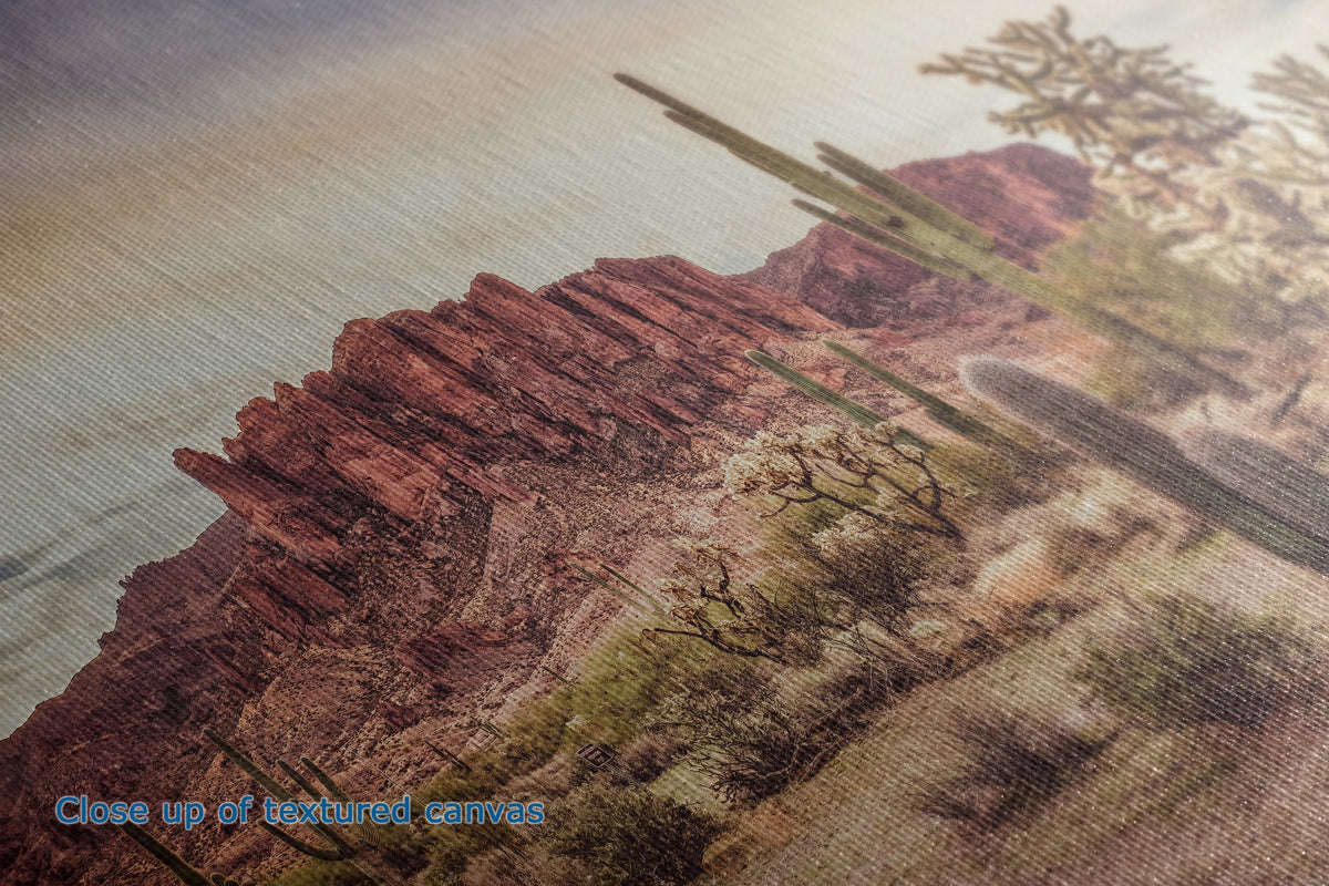 Close-up of the detail of a textured canvas print depicting a desert landscape with red rock cliffs, saguaro cacti, and a pale sky. Blue text in the lower-left corner reads 'Close up of textured canvas'.