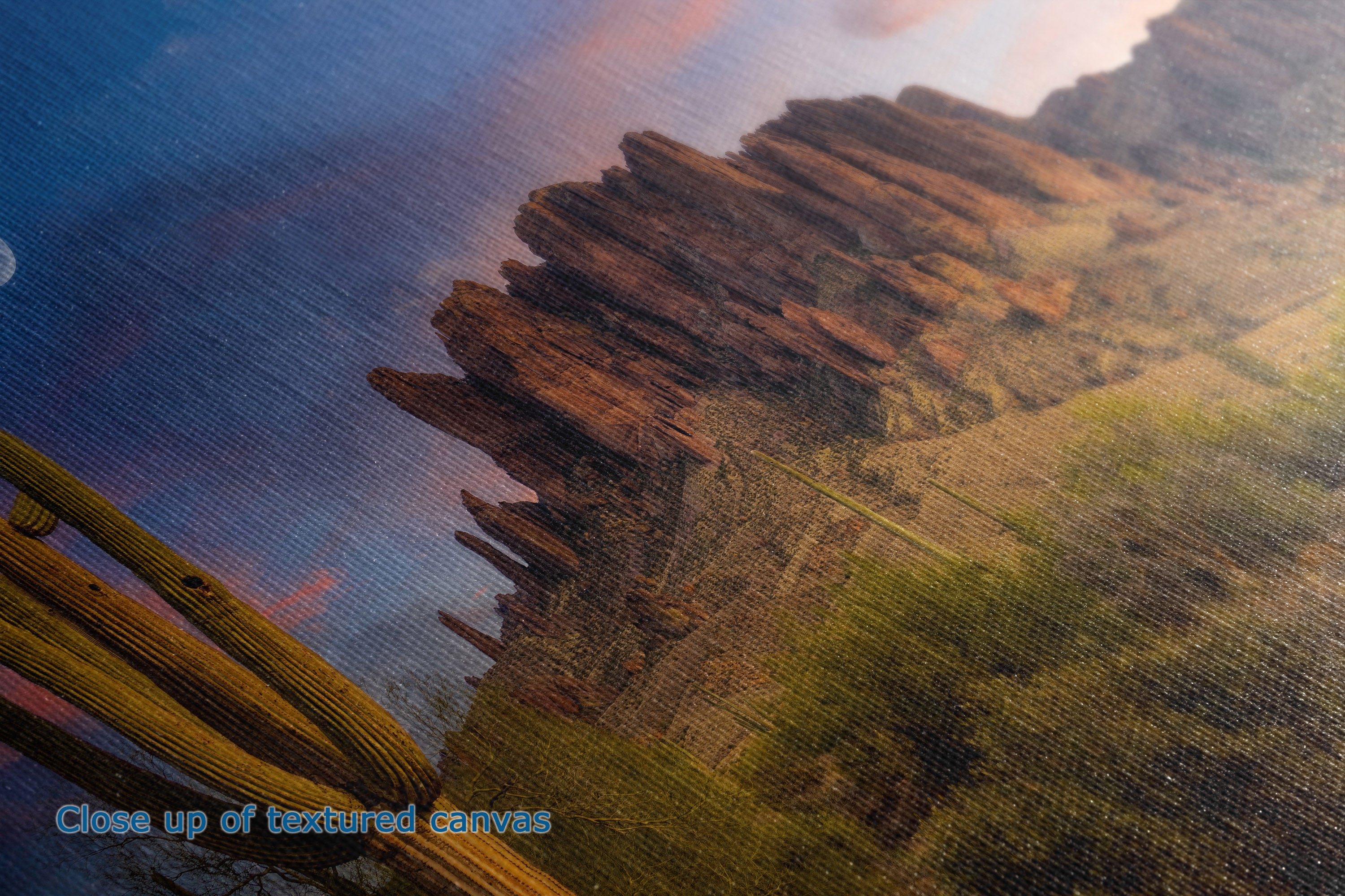 Close-up of a textured canvas print depicting a desert landscape with a tall reddish-brown rock cliff, green cacti, and a blue sky with warm sunset tones.