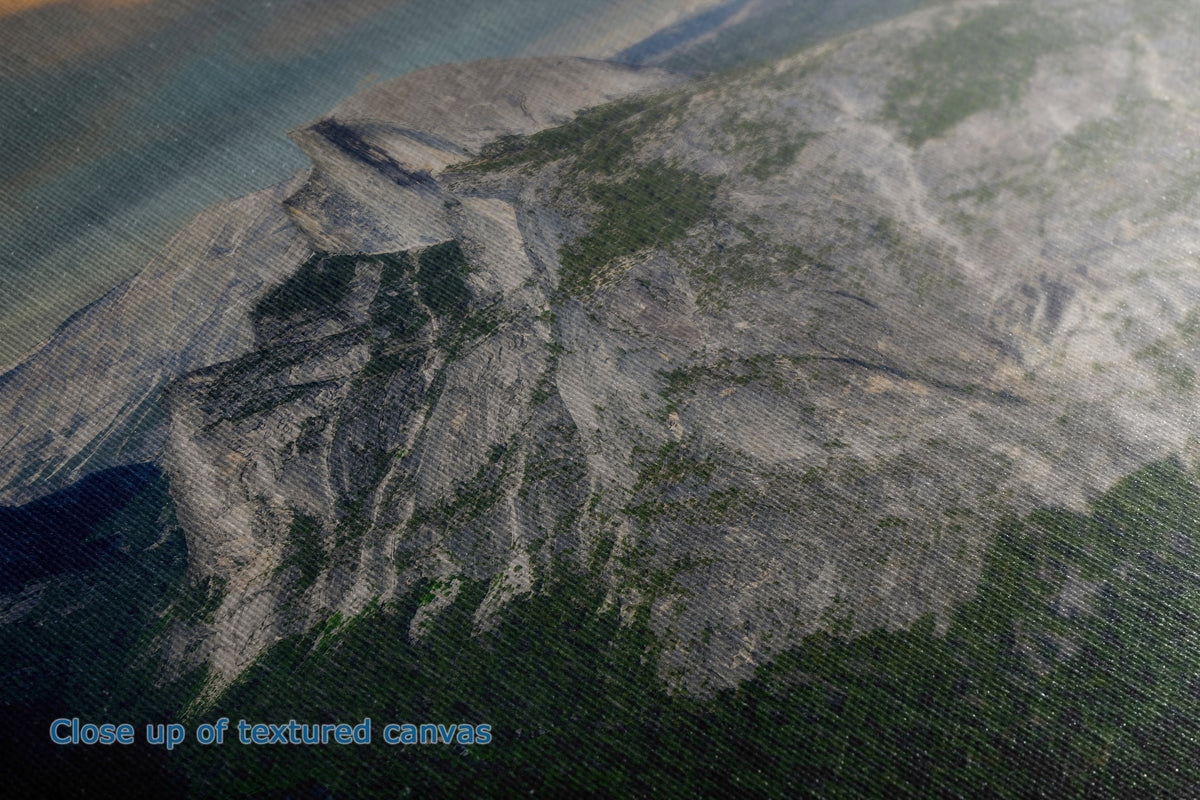 Close-up of a textured canvas print depicting a mountain landscape with steep rocky cliffs and dark green forested slopes under a hazy sky.