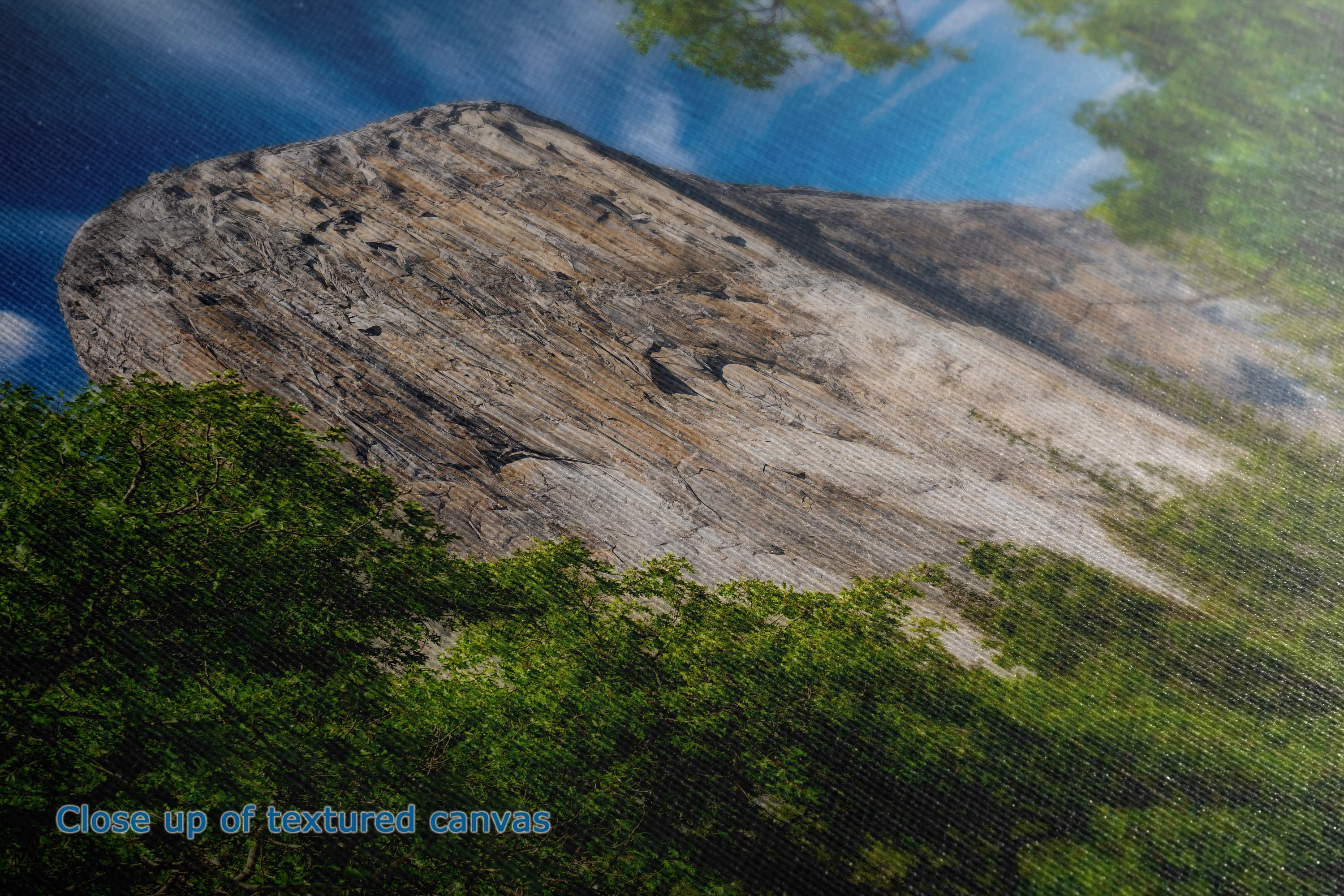 Close-up of the detail of a textured canvas print depicting a natural landscape with a large rock formation under a blue sky, framed by green trees. A small text overlay in the bottom-left corner reads 'Close up of textured canvas'.