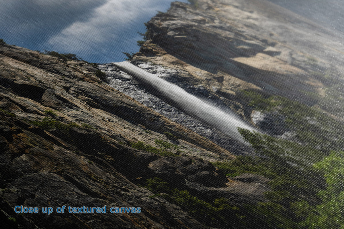 Close-up of the detail and texture of a canvas print depicting a dramatic waterfall cascading down a rugged rocky cliffside under a cloudy blue sky, with green vegetation in the foreground.