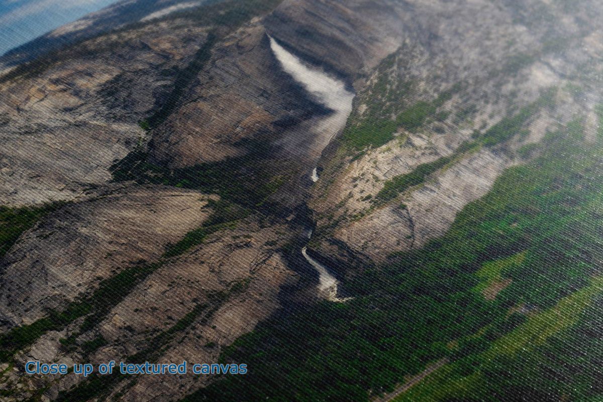 A close up detail and textured view of a canvas wall art print depicting a rugged mountain landscape with a winding river, green forests, and rocky cliffs.