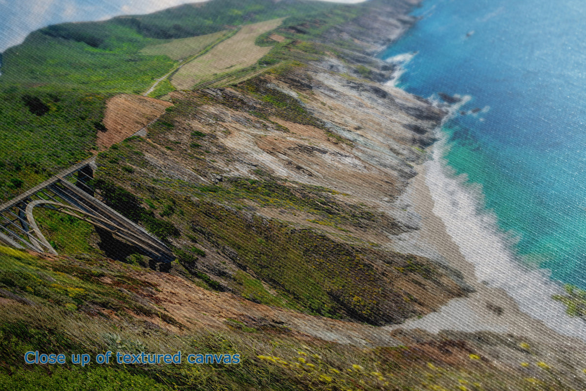 A close-up textured view of a coastal landscape with steep green hills, a winding road, rocky shoreline, and bright blue ocean water. Small blue text in the lower left reads 'Close up of textured canvas.