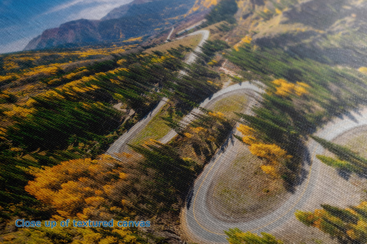 A close-up of the detail and texture of a canvas print depicting an aerial view of a winding mountain road surrounded by autumn-colored trees in yellow, green, and brown.