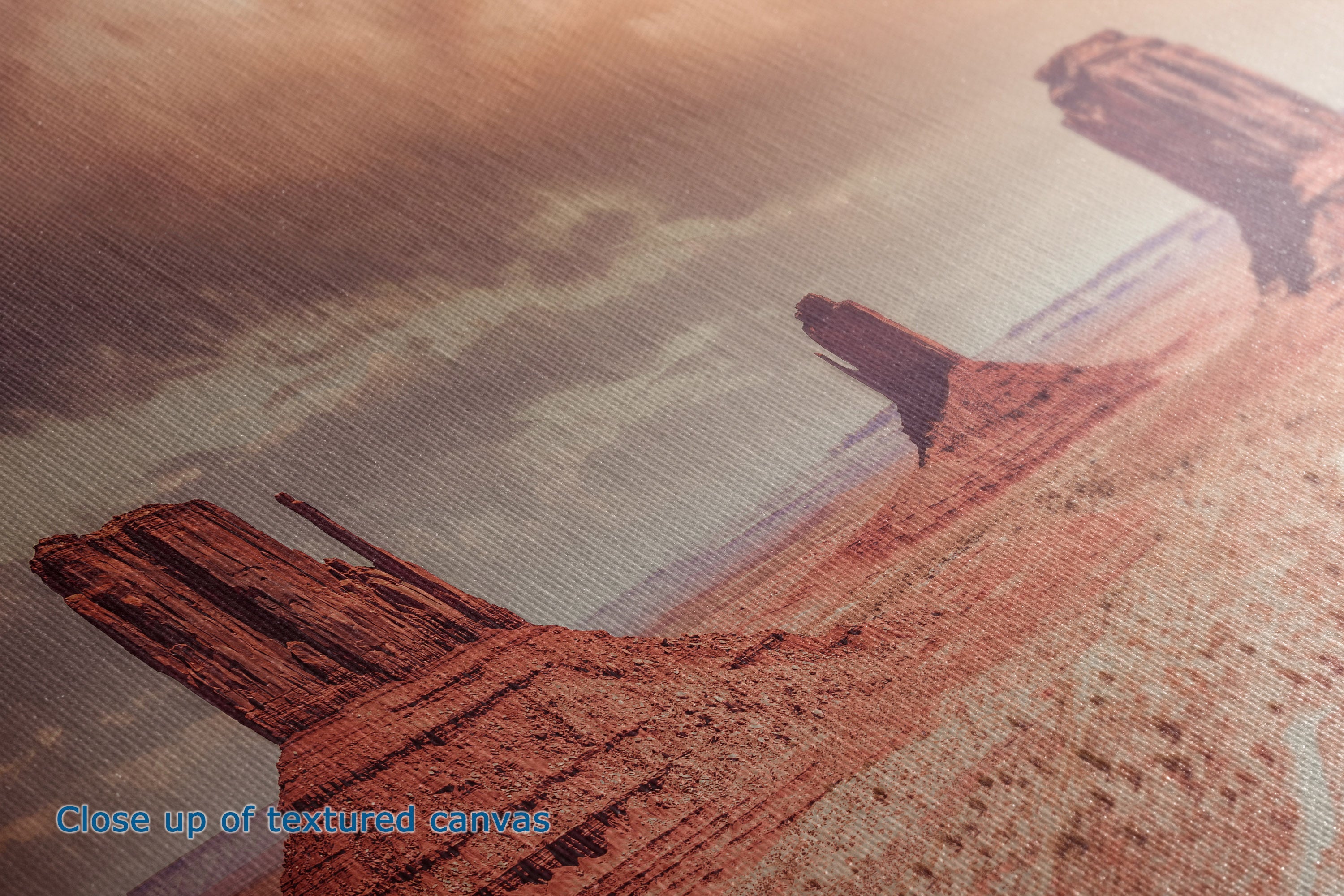 Close-up of a textured canvas print depicting a desert landscape with red rock buttes under a warm orange sky, with blue text reading 'Close up of textured canvas' in the lower left corner.