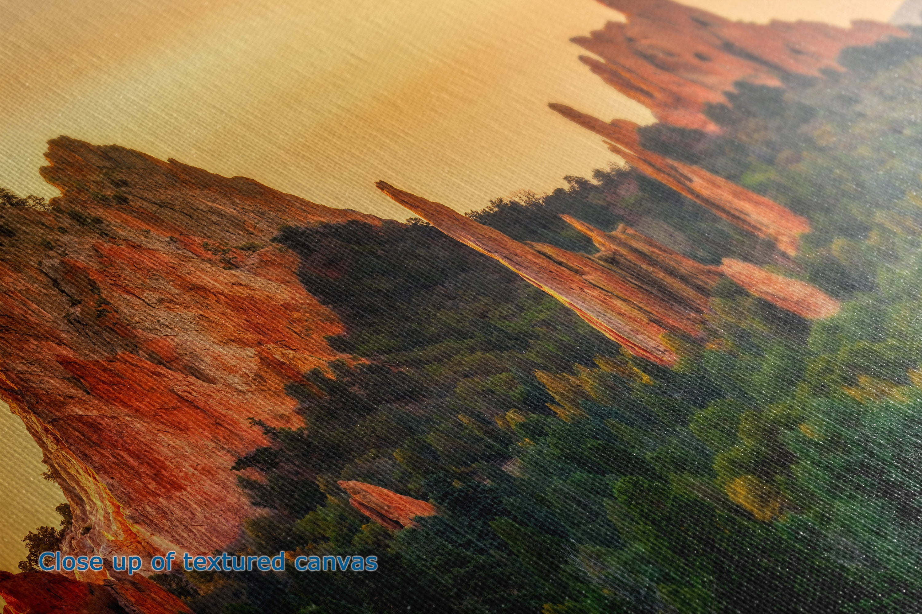 Close-up of the detail and texture of a canvas print depicting a landscape with reddish-brown rock formations and dark green forest under a warm golden sky.