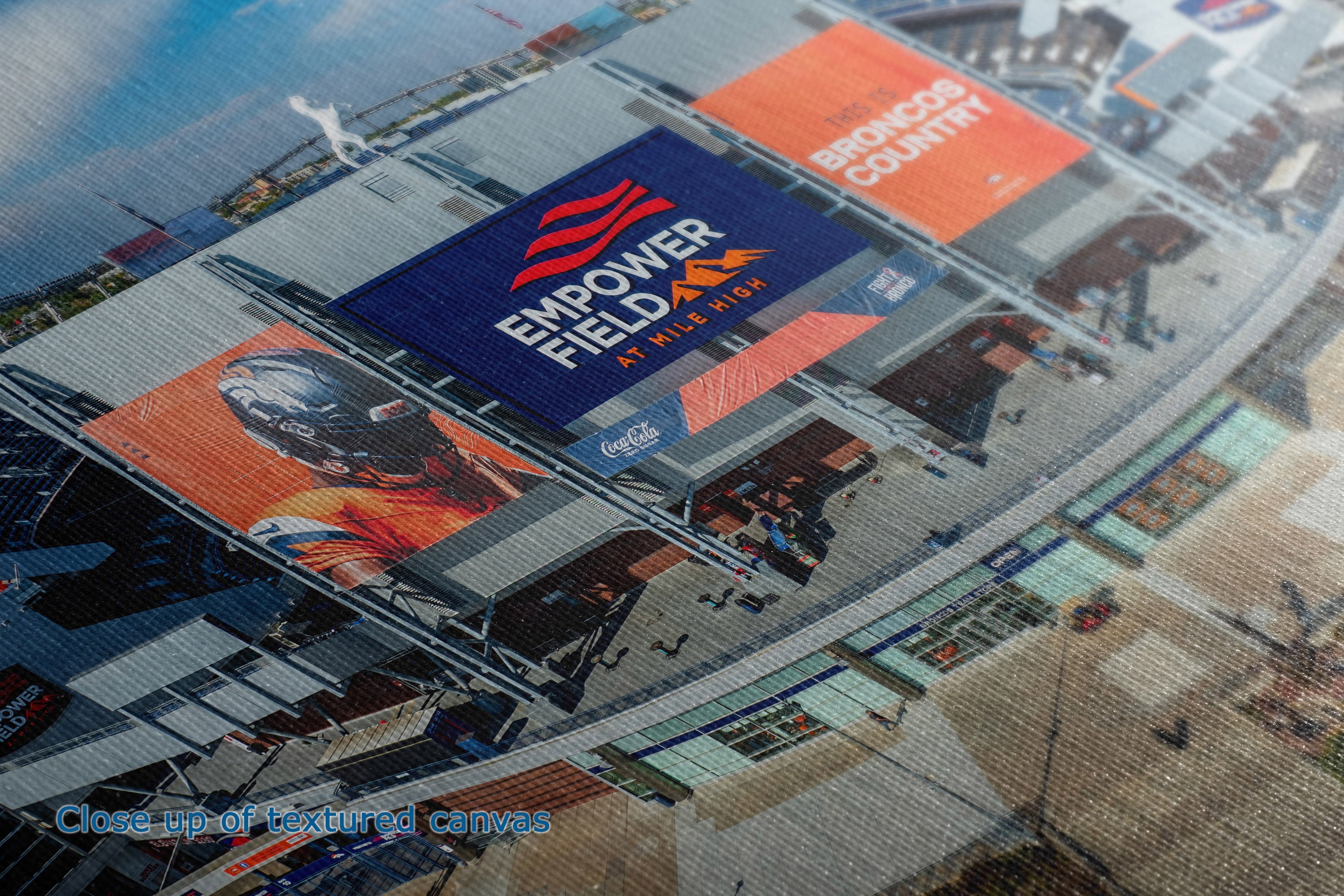 Close-up of the detail and texture of a canvas print depicting an aerial view of the Empower Field at Kilbish Park stadium, featuring large orange and blue advertising banners.